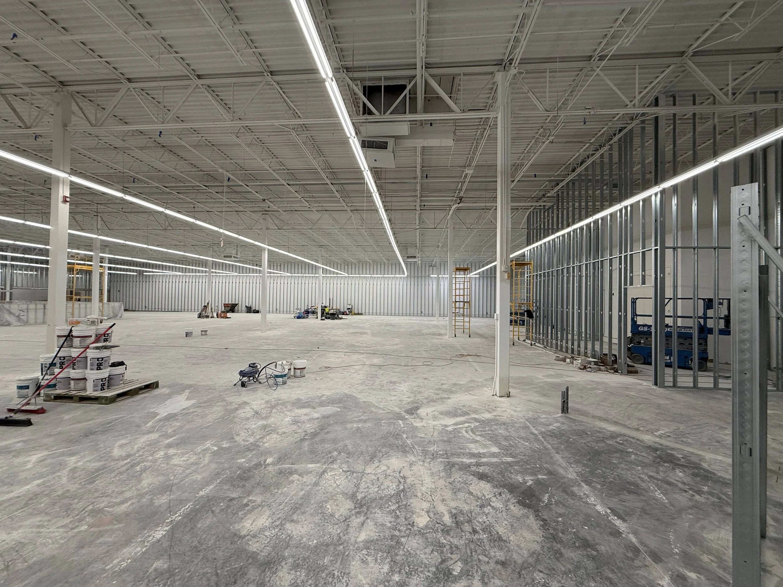 Large unfinished commercial or industrial building construction site with exposed metal framing, overhead lighting, and scattered construction tools and materials on the concrete floor.
