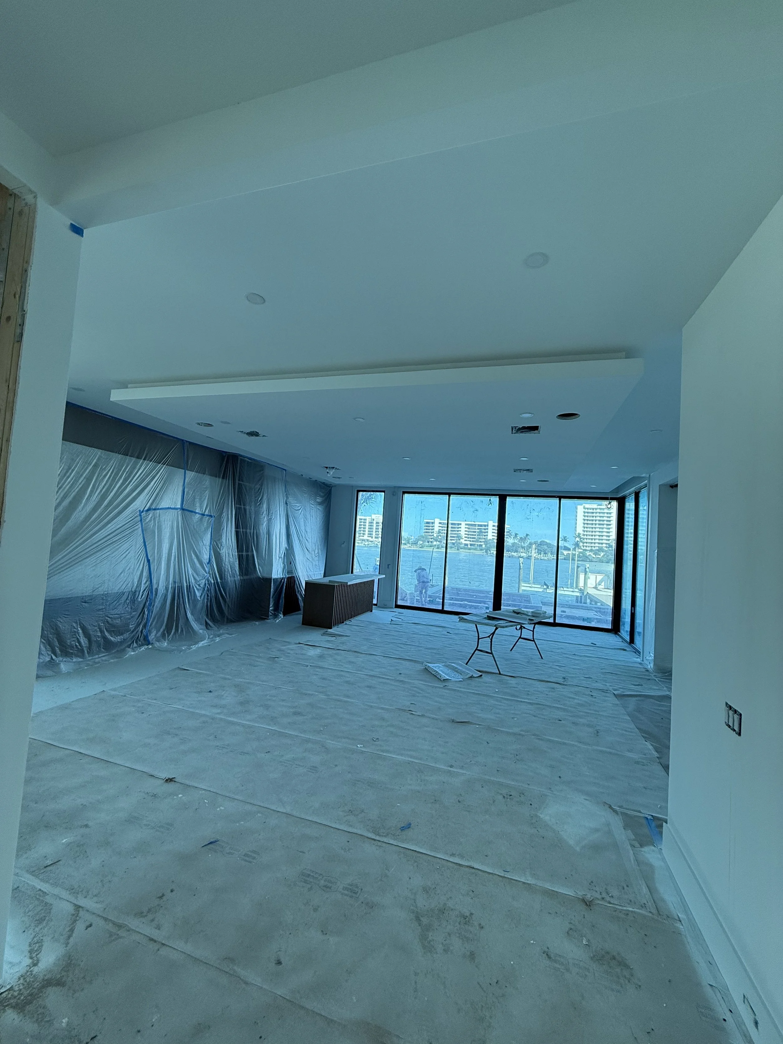 Living room under construction with plastic-covered furniture, large windows, and a ceiling with recessed lighting.