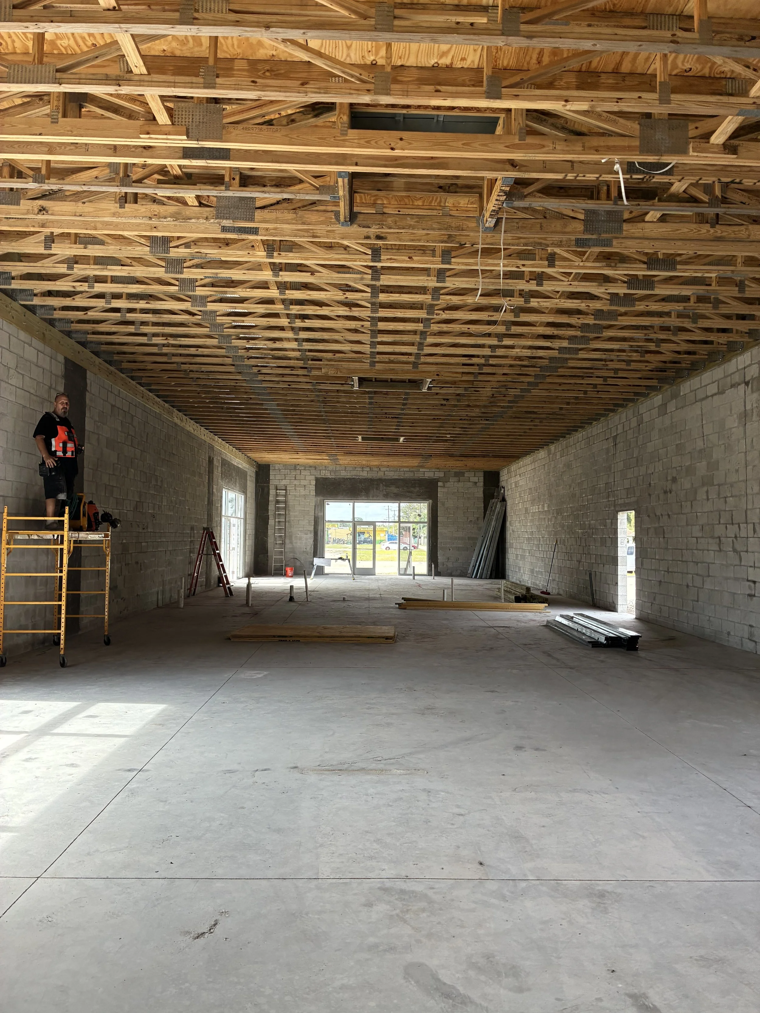 Interior of a building under construction with exposed wooden ceiling framework, unfinished concrete walls, and construction tools and materials on the floor, including a worker standing on a scaffold.