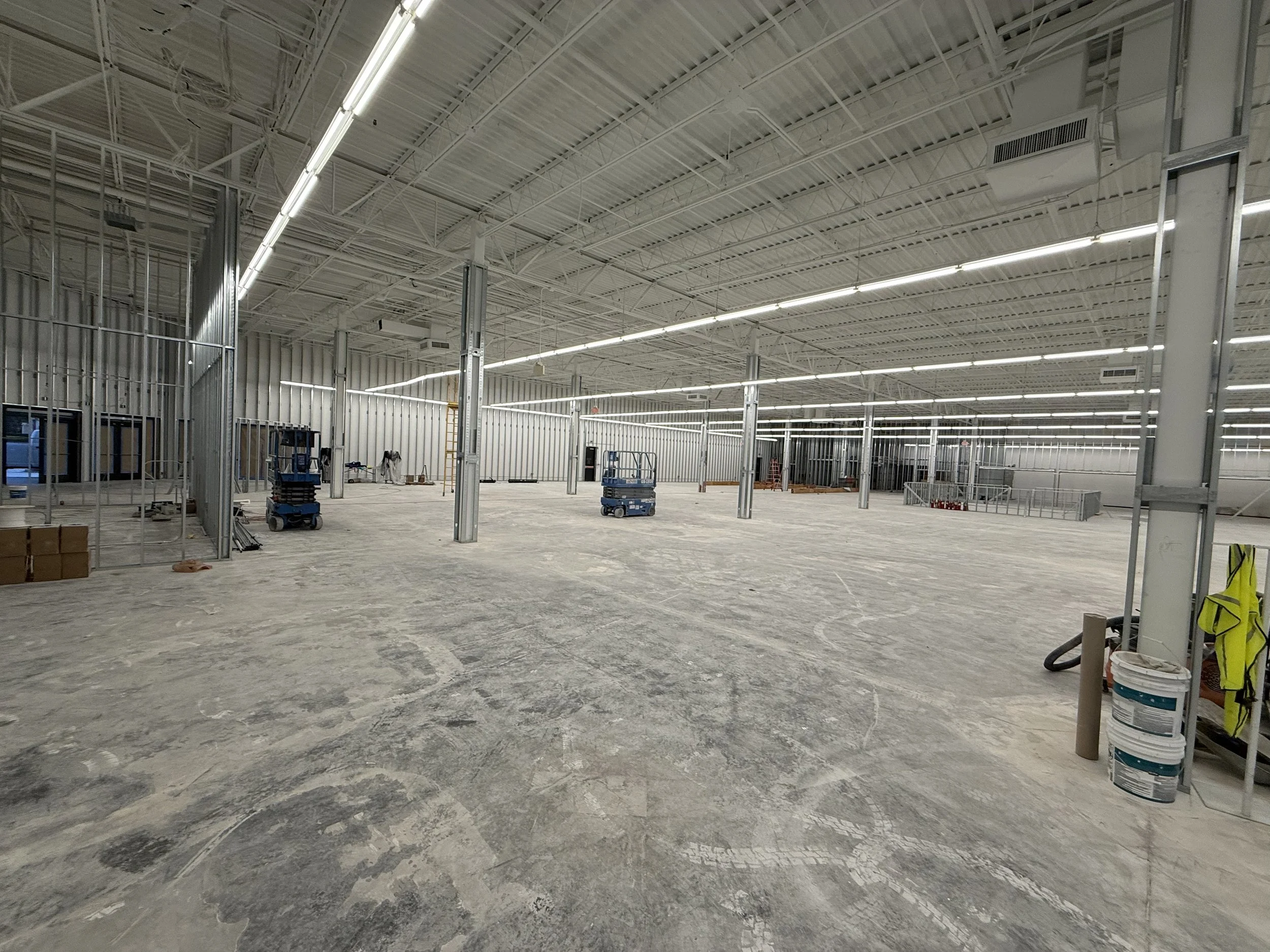 Interior of a large building under construction with exposed metal framework, unfinished concrete floor, and construction equipment.