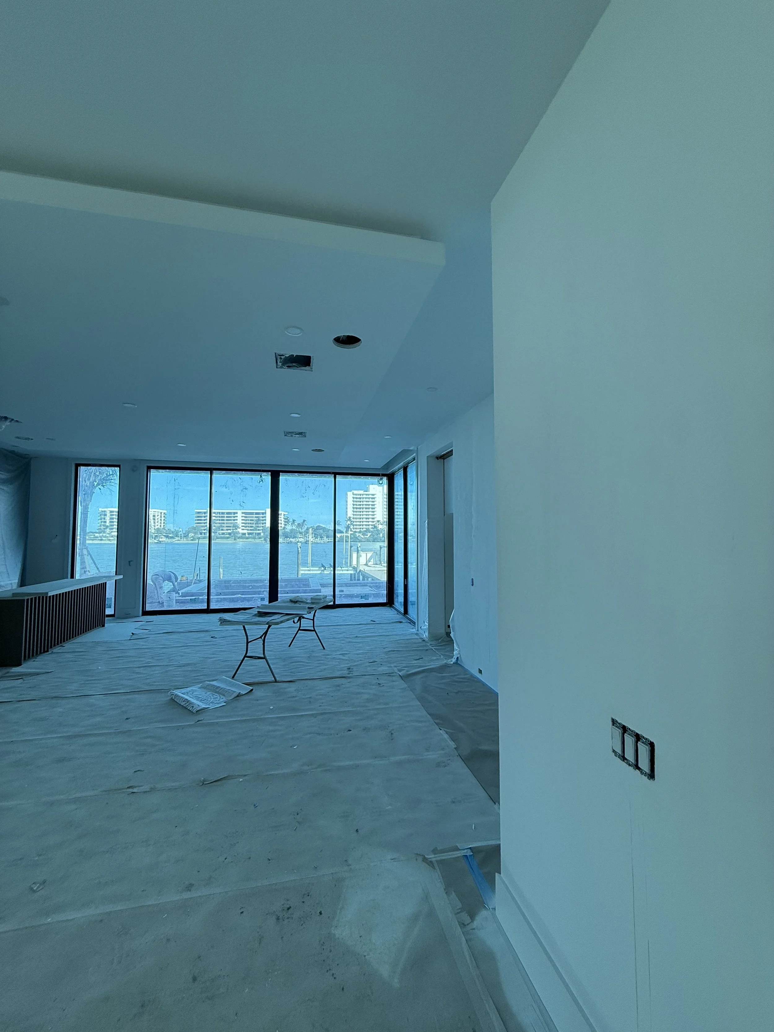 Interior of a room under construction with large windows overlooking a body of water, with plastic sheets on the floor and electrical outlets on the wall.