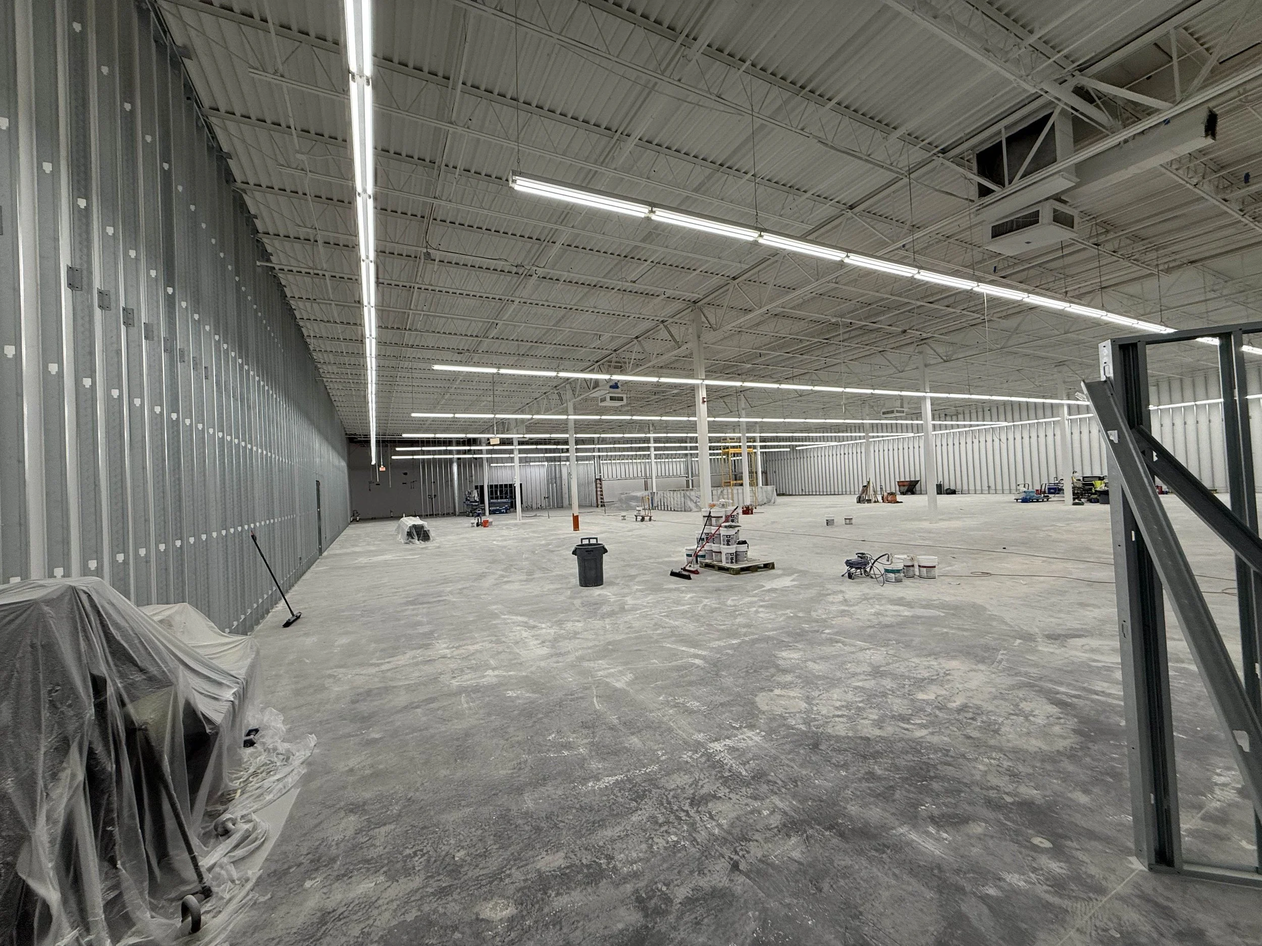 Large indoor construction space with exposed metal walls and ceiling, bright overhead lighting, construction tools, buckets, a ladder, and covered equipment, indicating ongoing building or renovation.