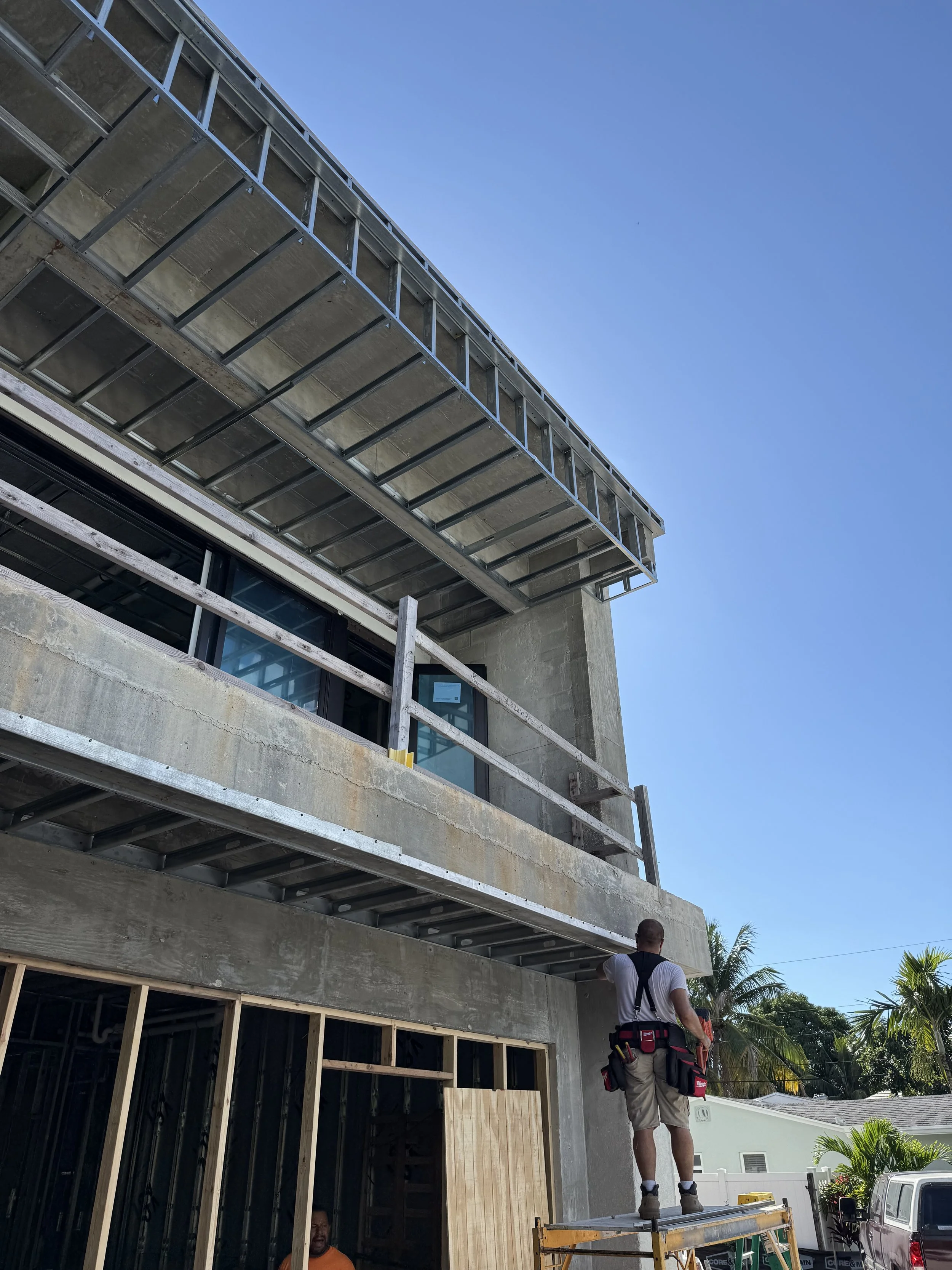 Construction worker standing on scaffolding on a building under construction, with support tools hanging from his tool belt, in front of unfinished concrete and window frames, while a bright blue sky and some palm trees are visible in the background.