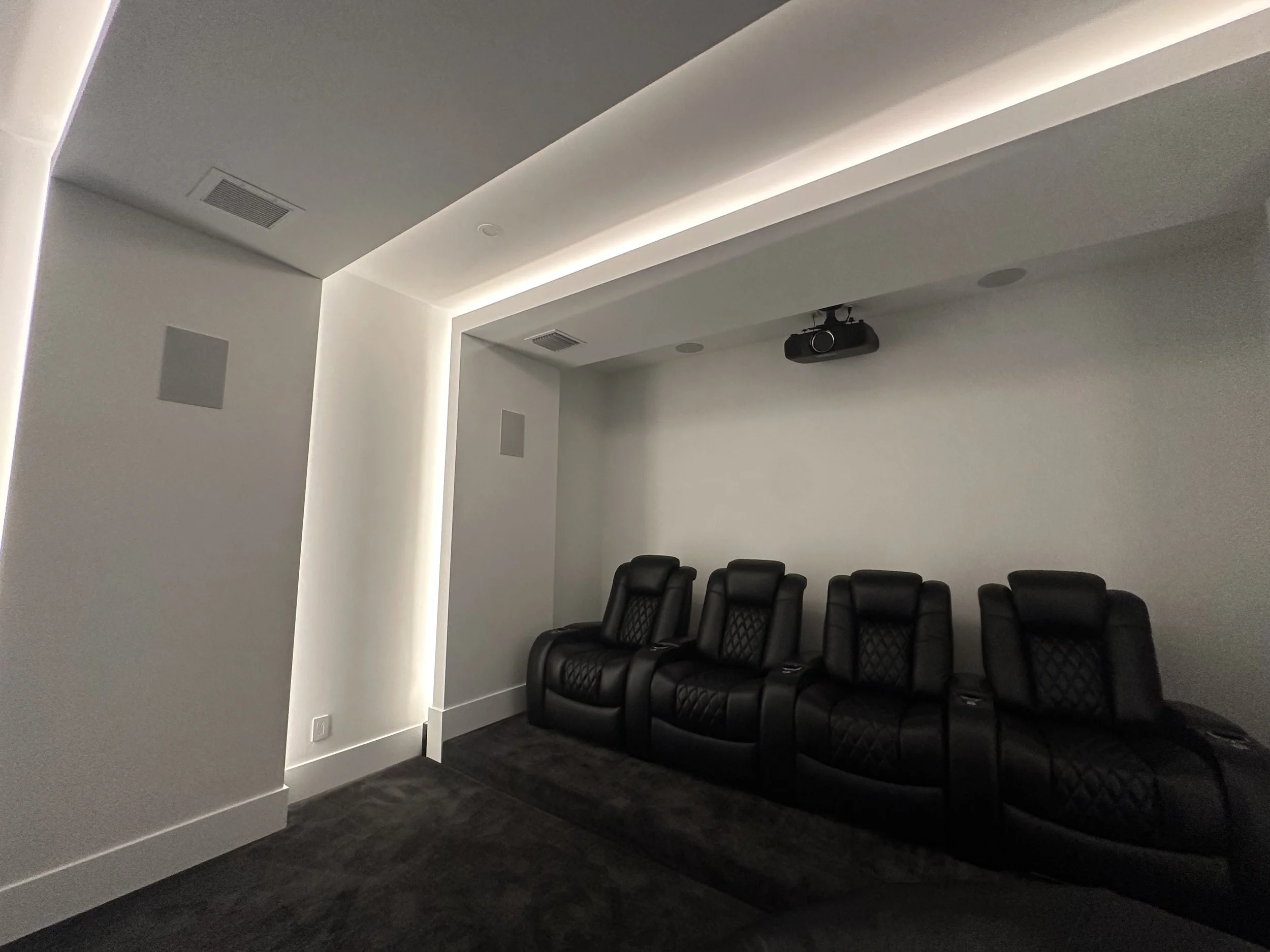 A modern home theater room with a row of five black leather recliner chairs, a ceiling-mounted projector, and a white wall with indirect lighting.