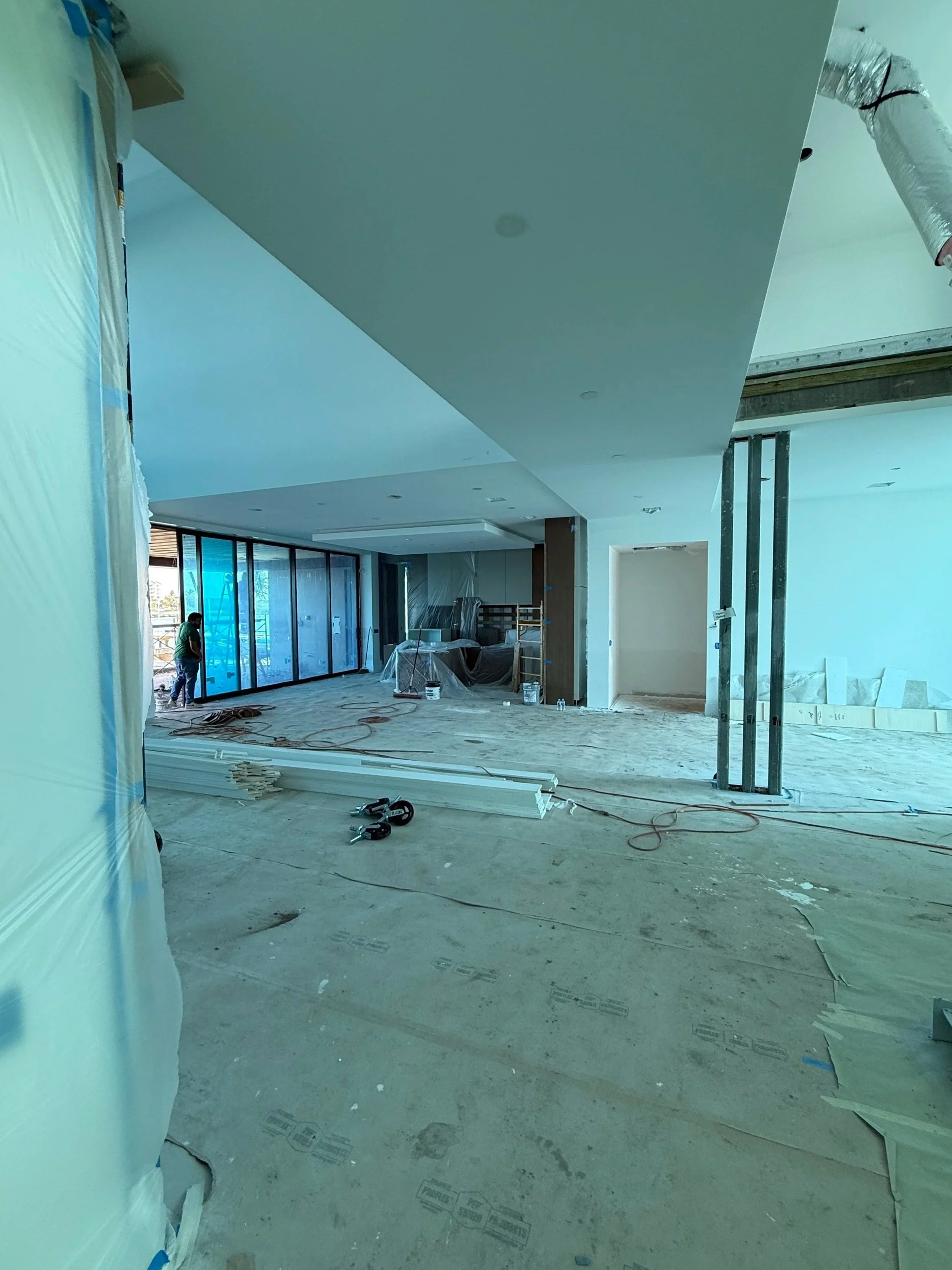 Interior of a building under construction or renovation, with construction tools, pipes, and materials scattered on the floor, some wrapped in plastic, and a worker in the background.