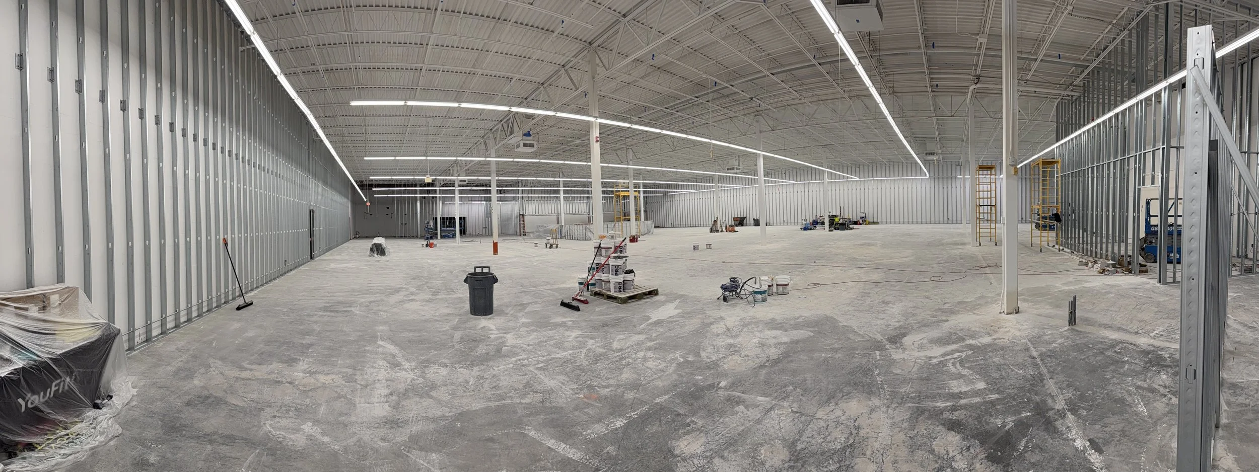 Interior of a large commercial building under construction with metal framing, exposed ceiling, and unfinished concrete floor. Construction tools and materials are scattered throughout the space.