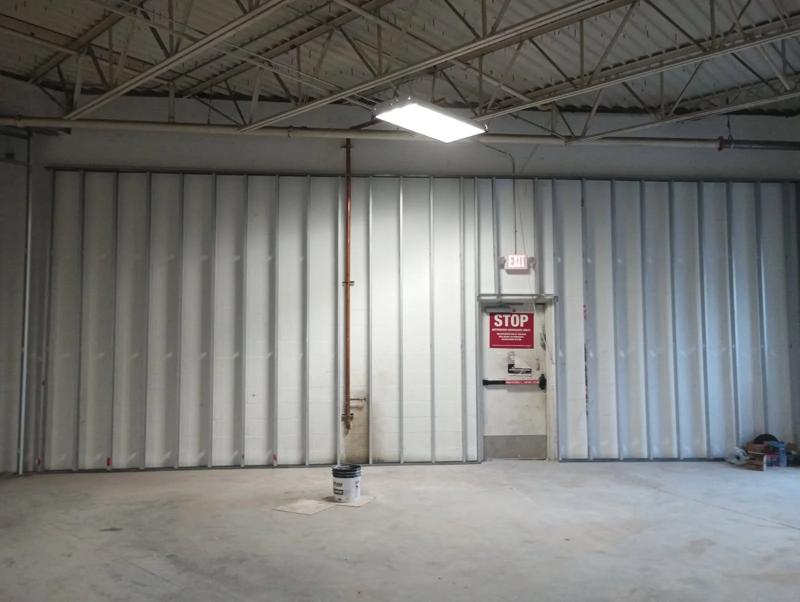 Interior of a construction site with unfinished walls, a pipe running vertically, a bucket on the floor, a red stop sign on a door, and an EXIT sign above the door.