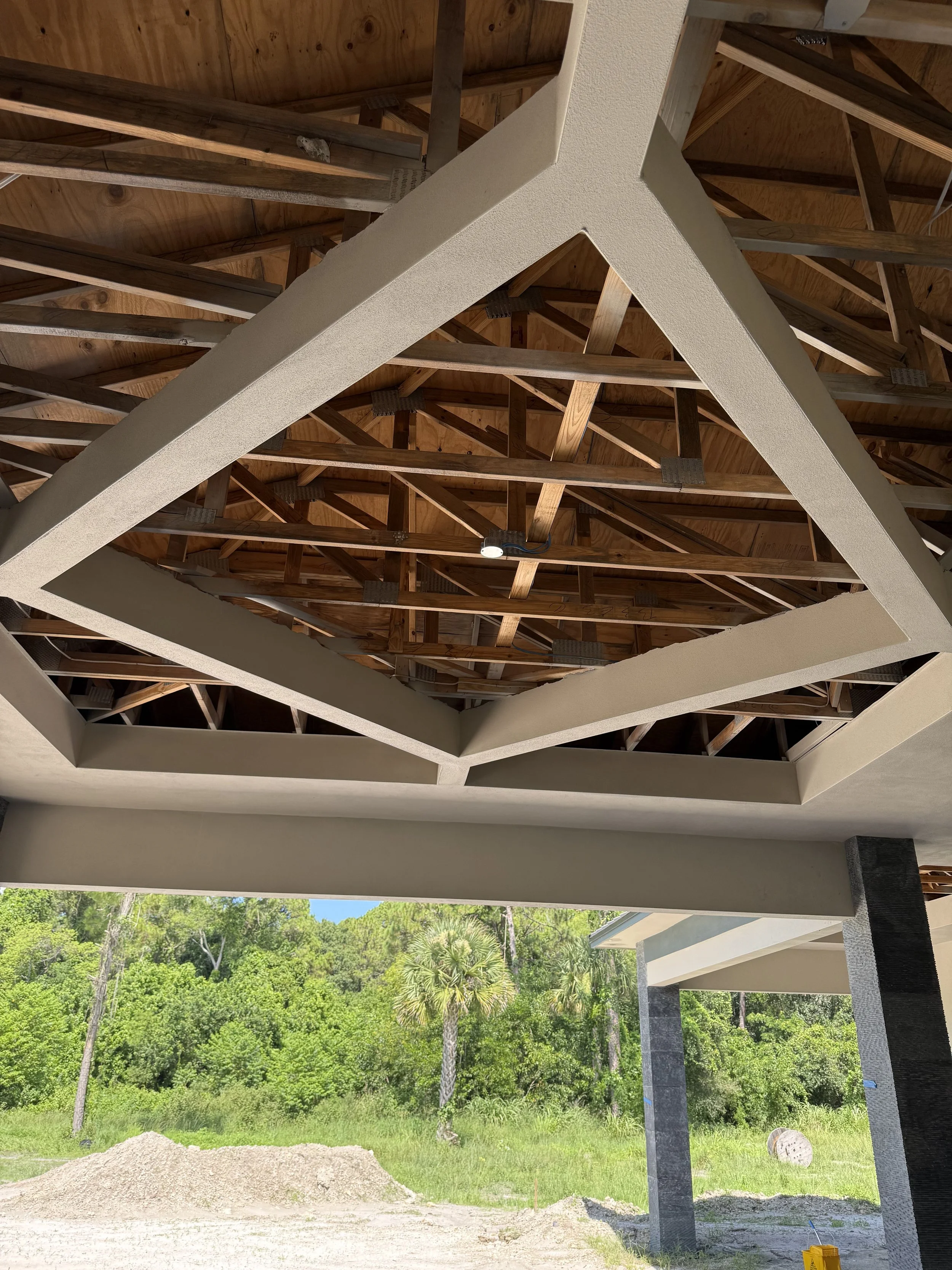 View of a construction site showing the underside of a building with a wooden roof structure and an unfinished exterior facing outdoors, surrounded by trees, with construction materials and dirt ground.