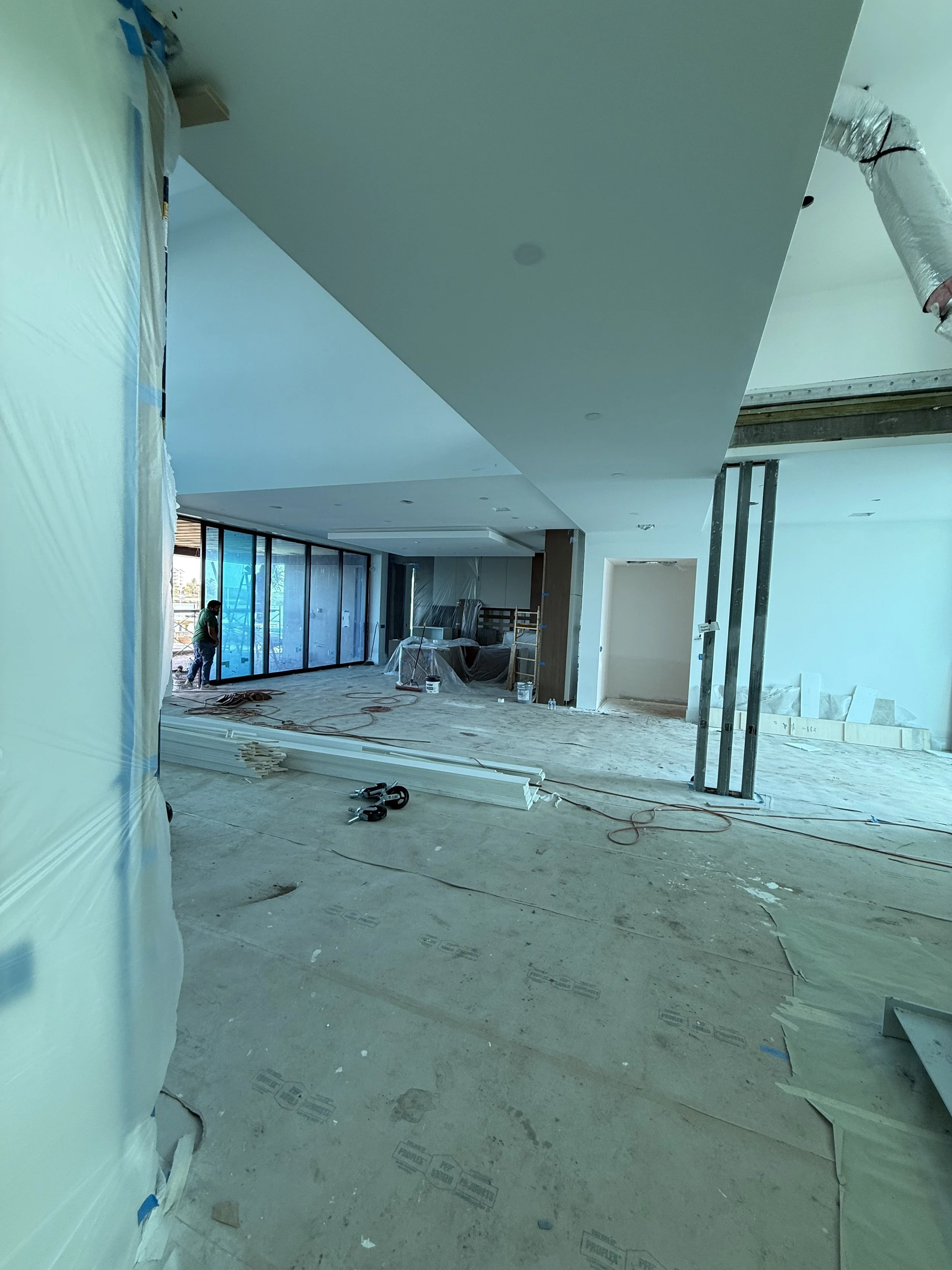 Interior of a house under construction with unfinished flooring, tools, and construction materials. Large windows along one side, and visible ductwork on the ceiling.