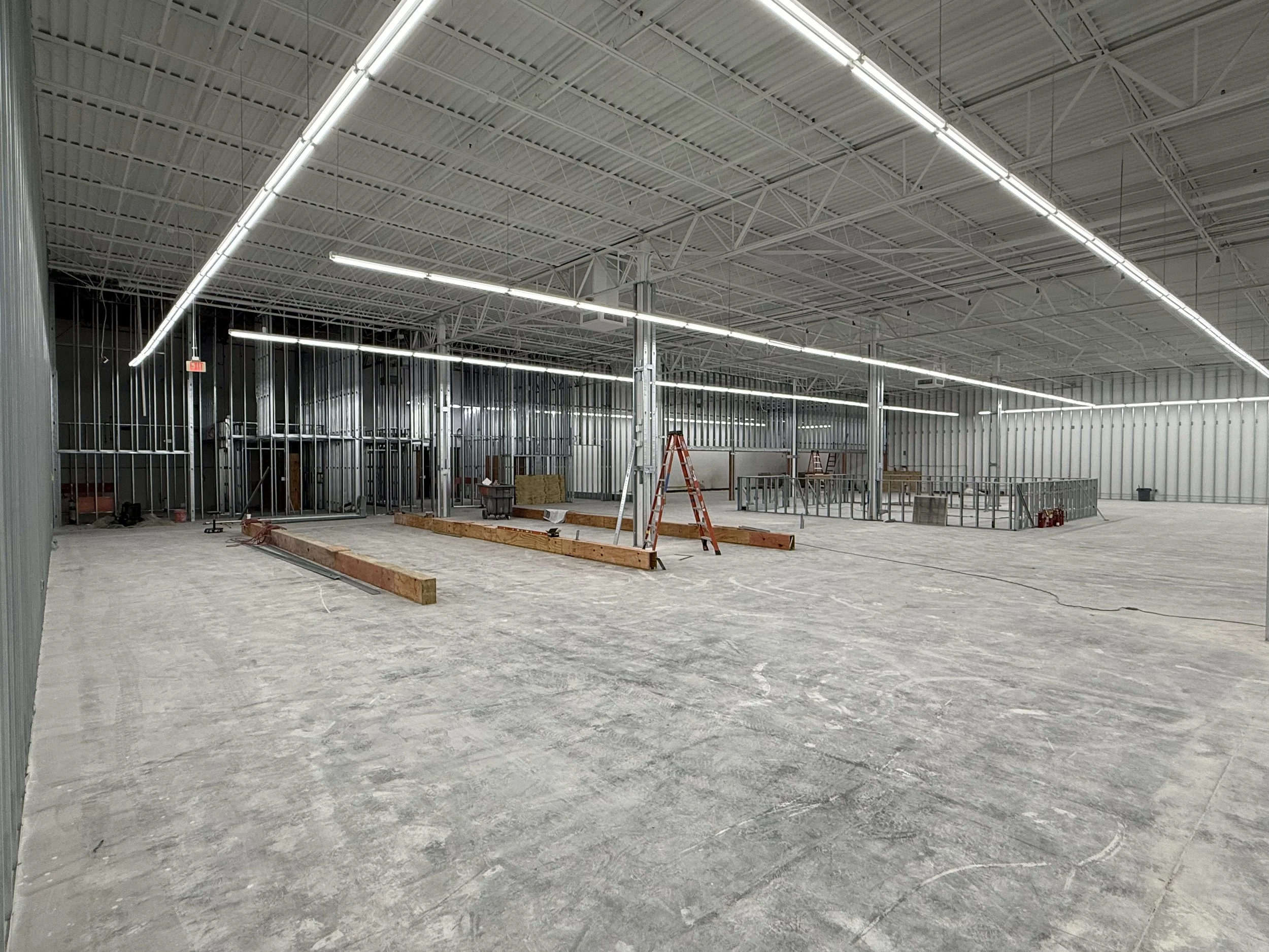Interior of a building under construction with metal framing, unfinished concrete floor, and exposed ceiling structure; construction materials and tools present.