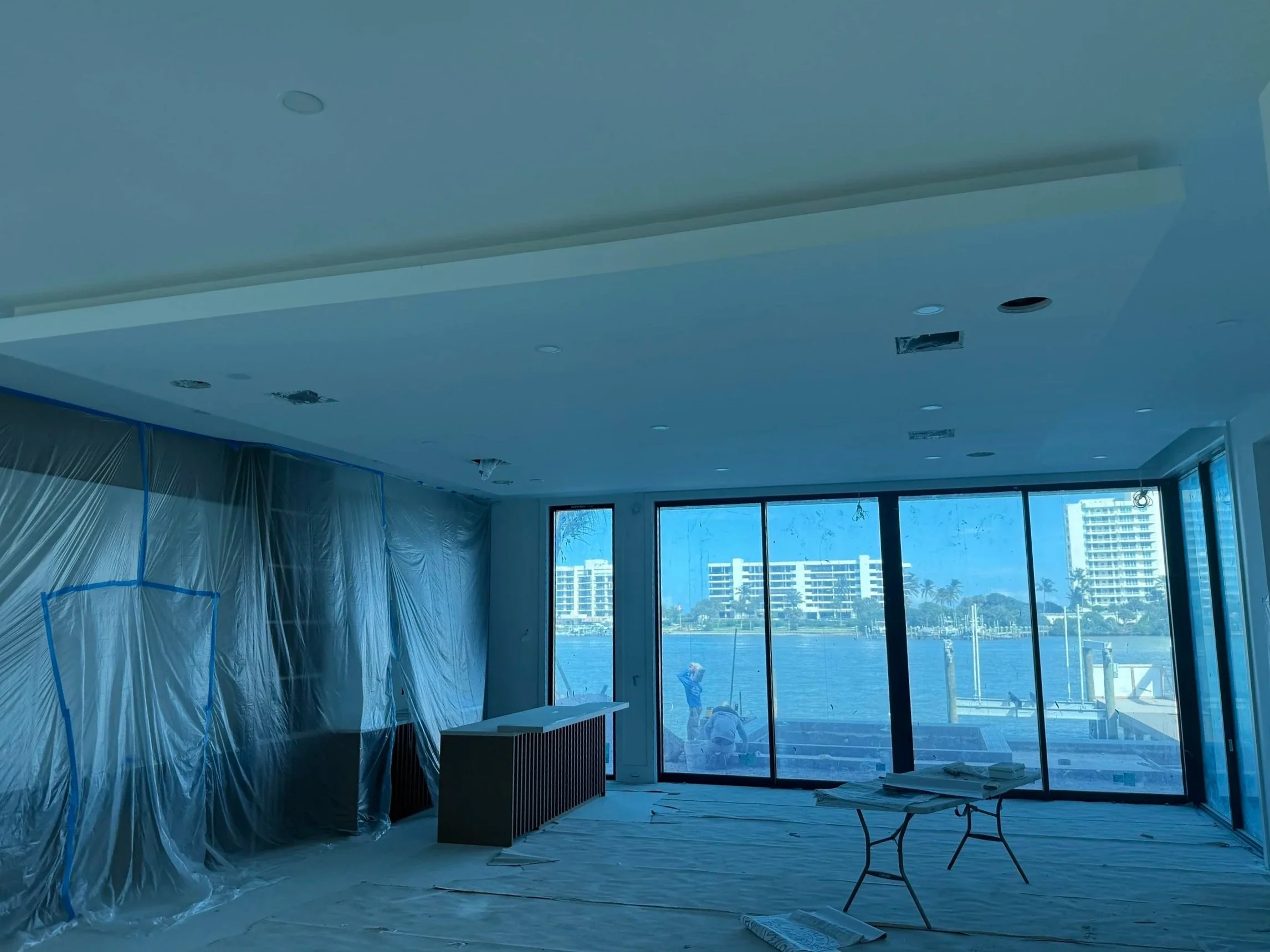 Interior of a room under construction with plastic sheeting covering the walls, a large floor-to-ceiling window showing a waterfront view with buildings and palm trees, and construction materials and tools on the floor.