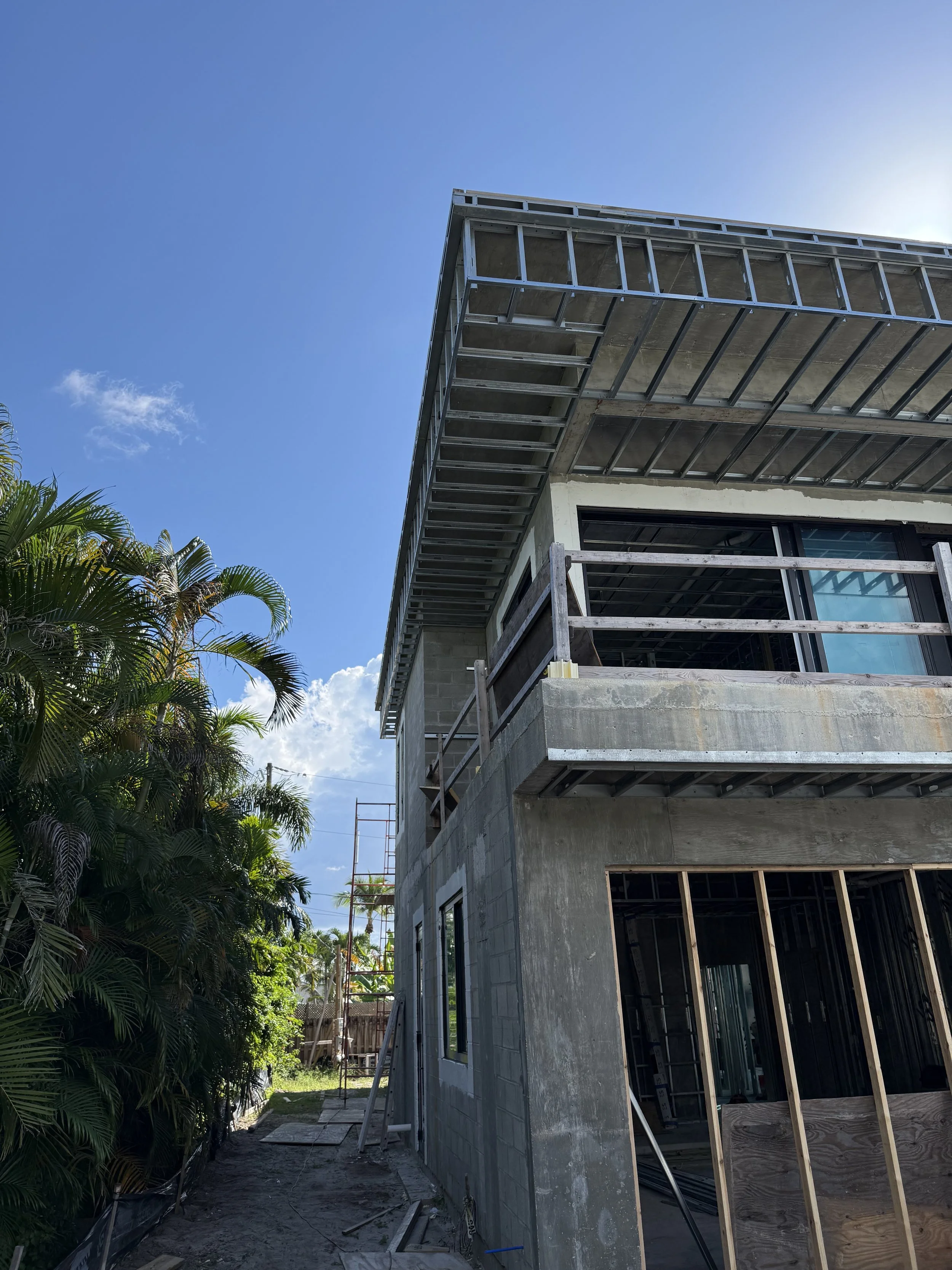 Construction site of a multi-story building with scaffolding and exterior work ongoing, surrounded by tropical plants and a clear blue sky.
