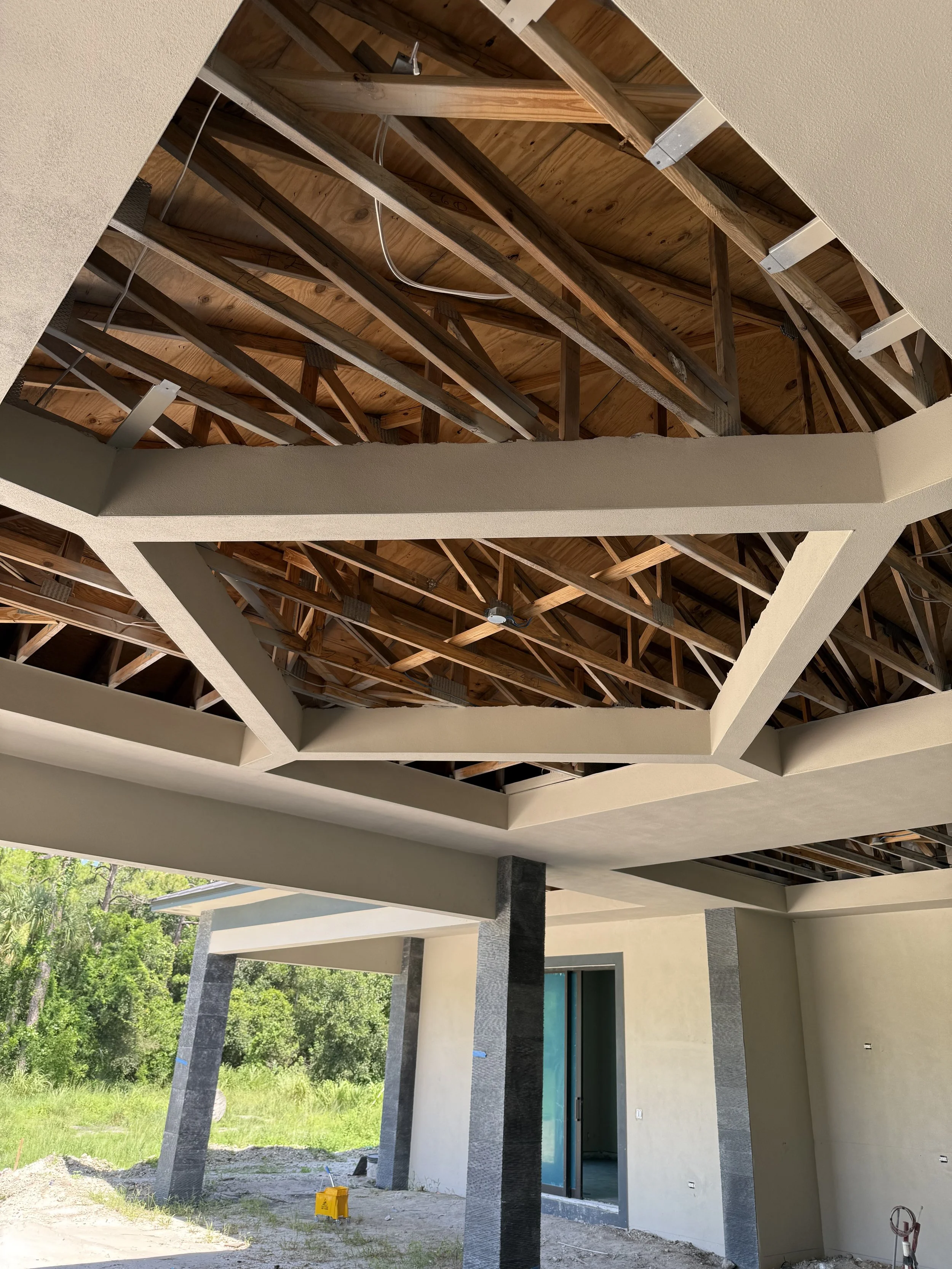 Construction site of a house with a partially built ceiling showing exposed wooden beams and metal supports, with a covered patio area and a doorway leading inside, surrounded by greenery.