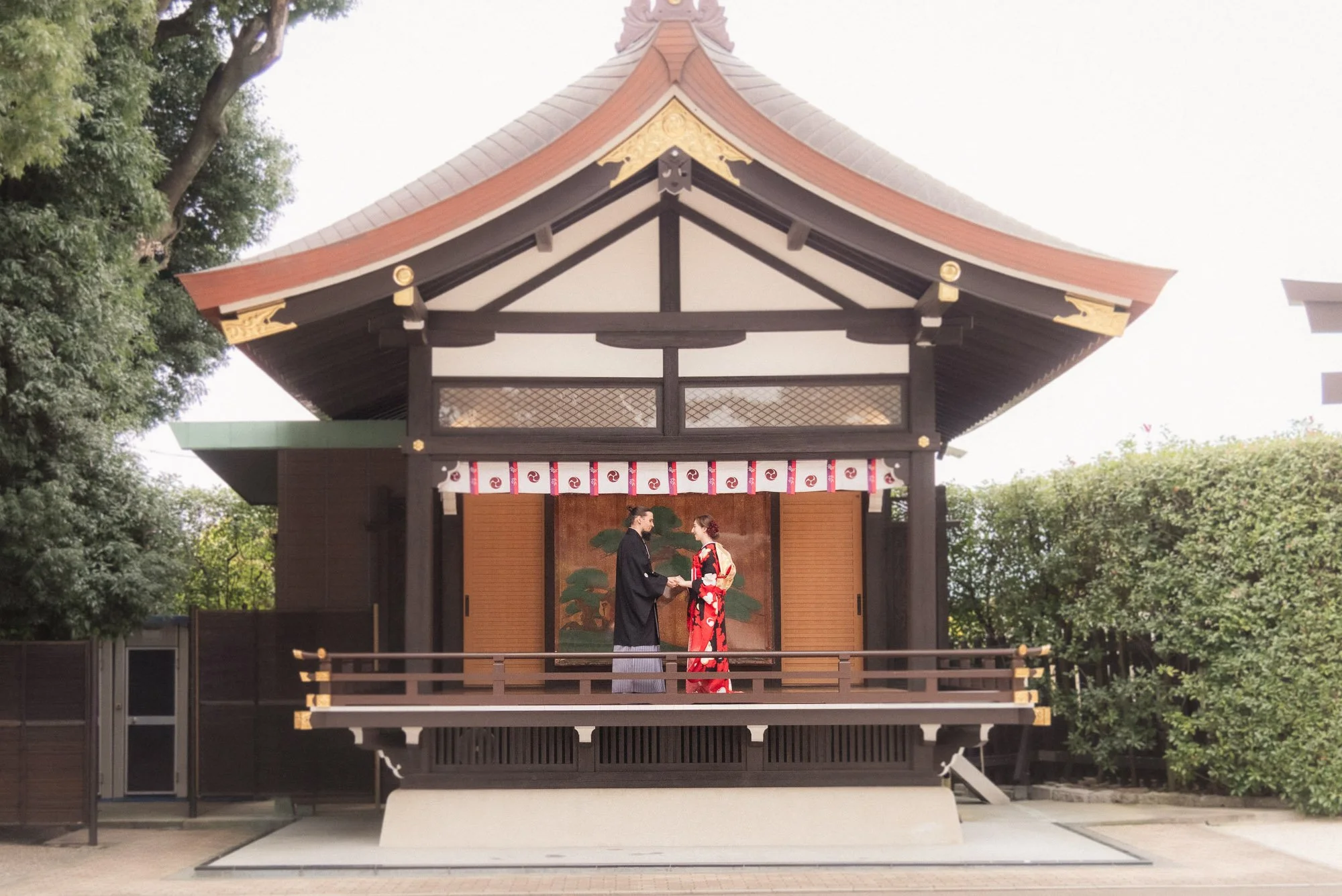 Couple wearing wedding kimonos stand on a noh theatre within the grounds of a beautiful shrine in Japan.