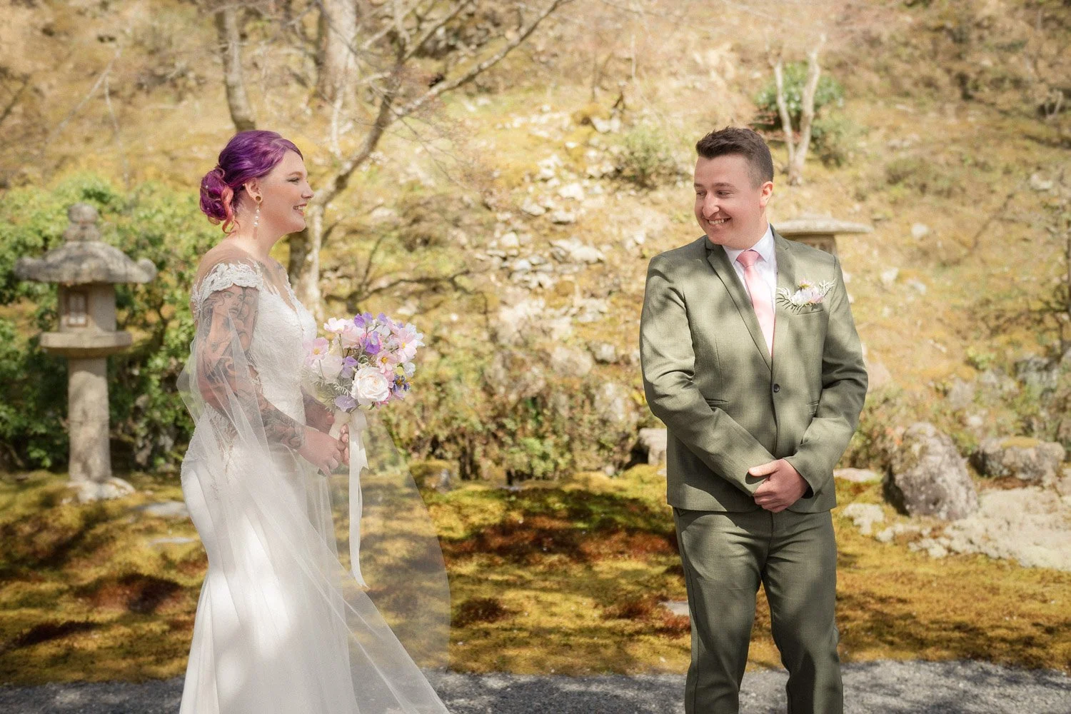 A bride and groom smiling at each other during a first reveal in a Japanese garden, with moss, stone lanterns, and early spring foliage in the background.