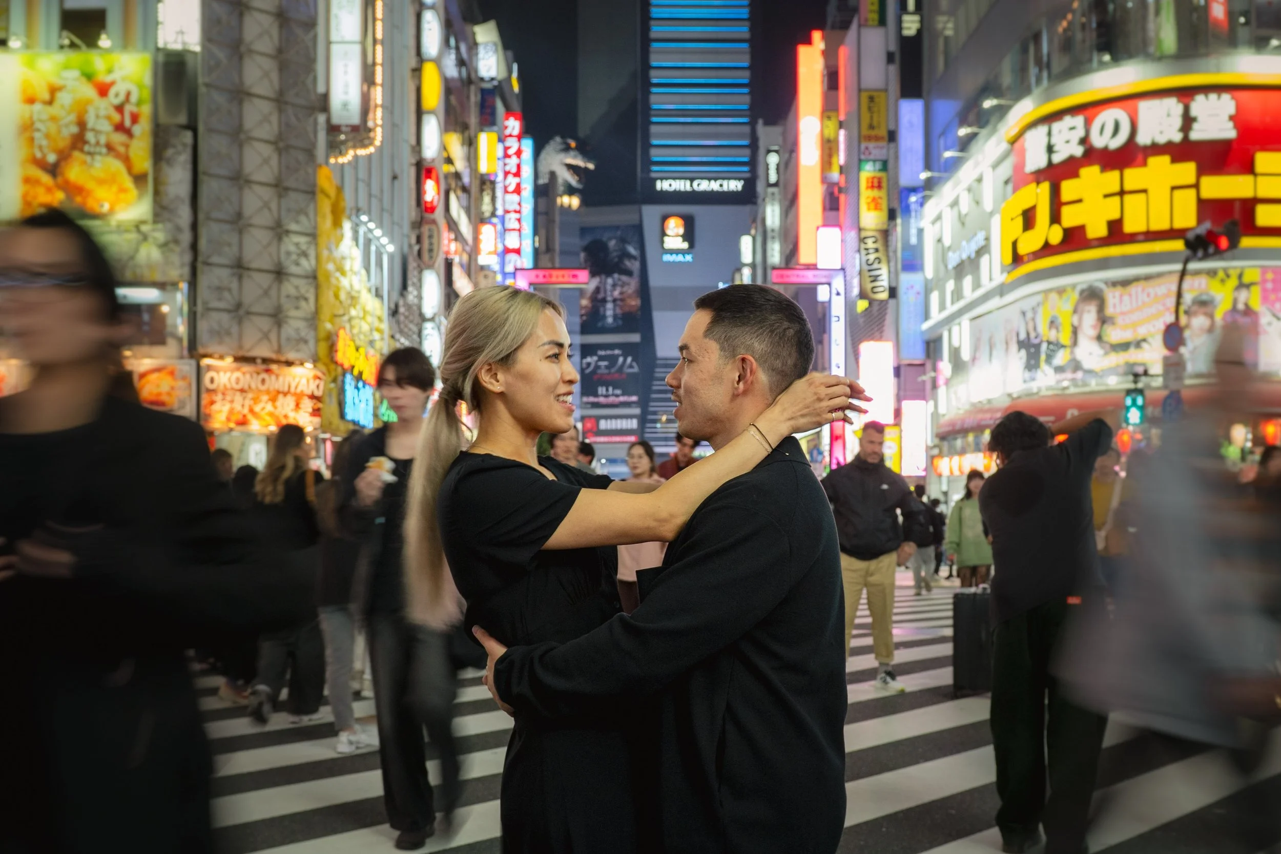 A couple is dancing closely together in the middle of a busy city street at night, surrounded by illuminated signs and crowds.