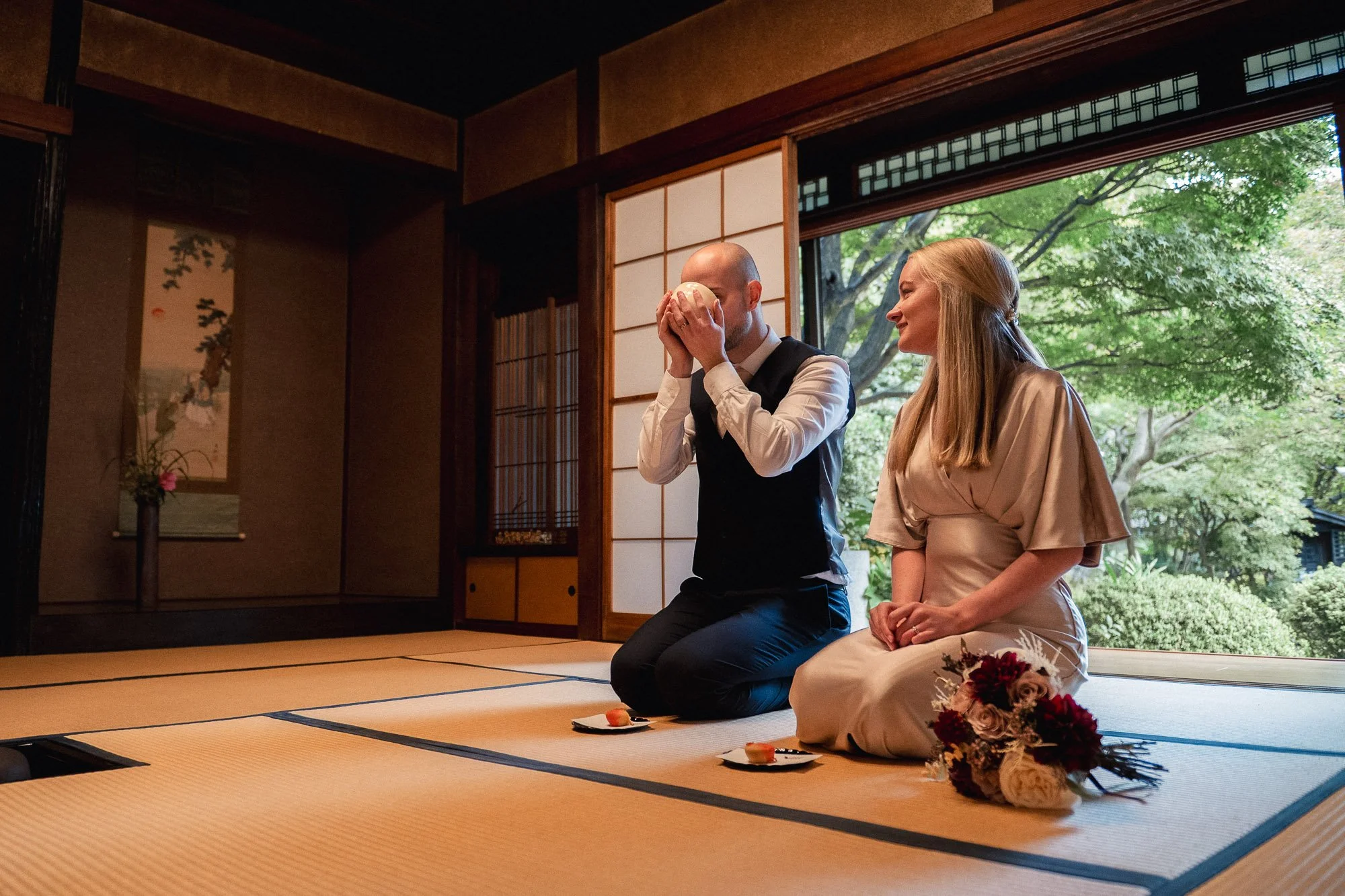 A wedding ceremony with a man and woman kneeling on tatami mats inside a traditional Japanese room with shoji screens, outdoors view of green trees, man holding a sake cup, woman with a bouquet