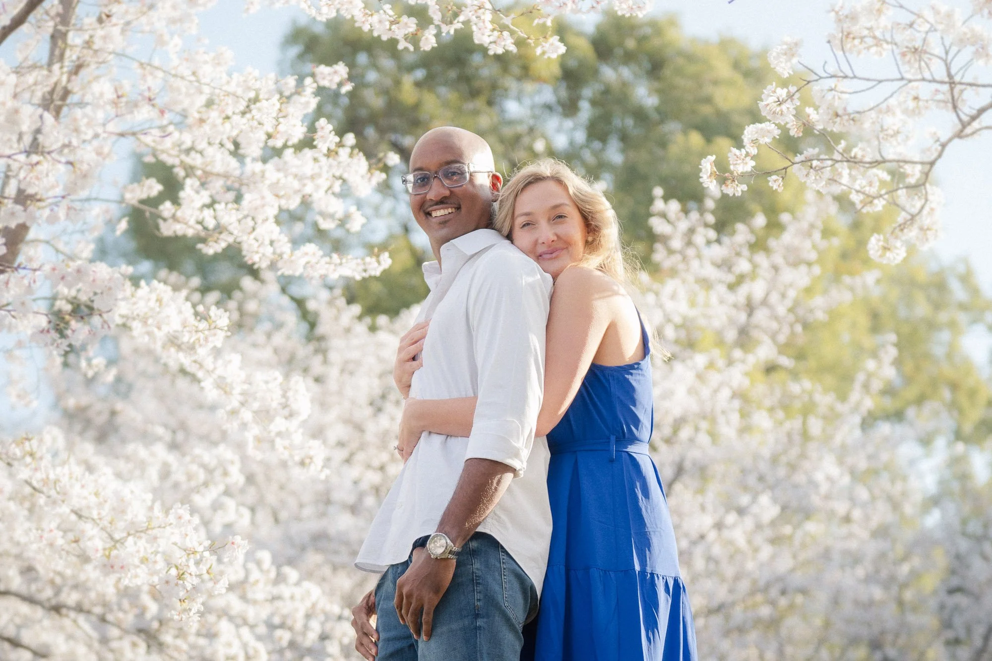 A smiling couple standing close together outdoors amidst blooming white cherry blossom trees on a sunny day.
