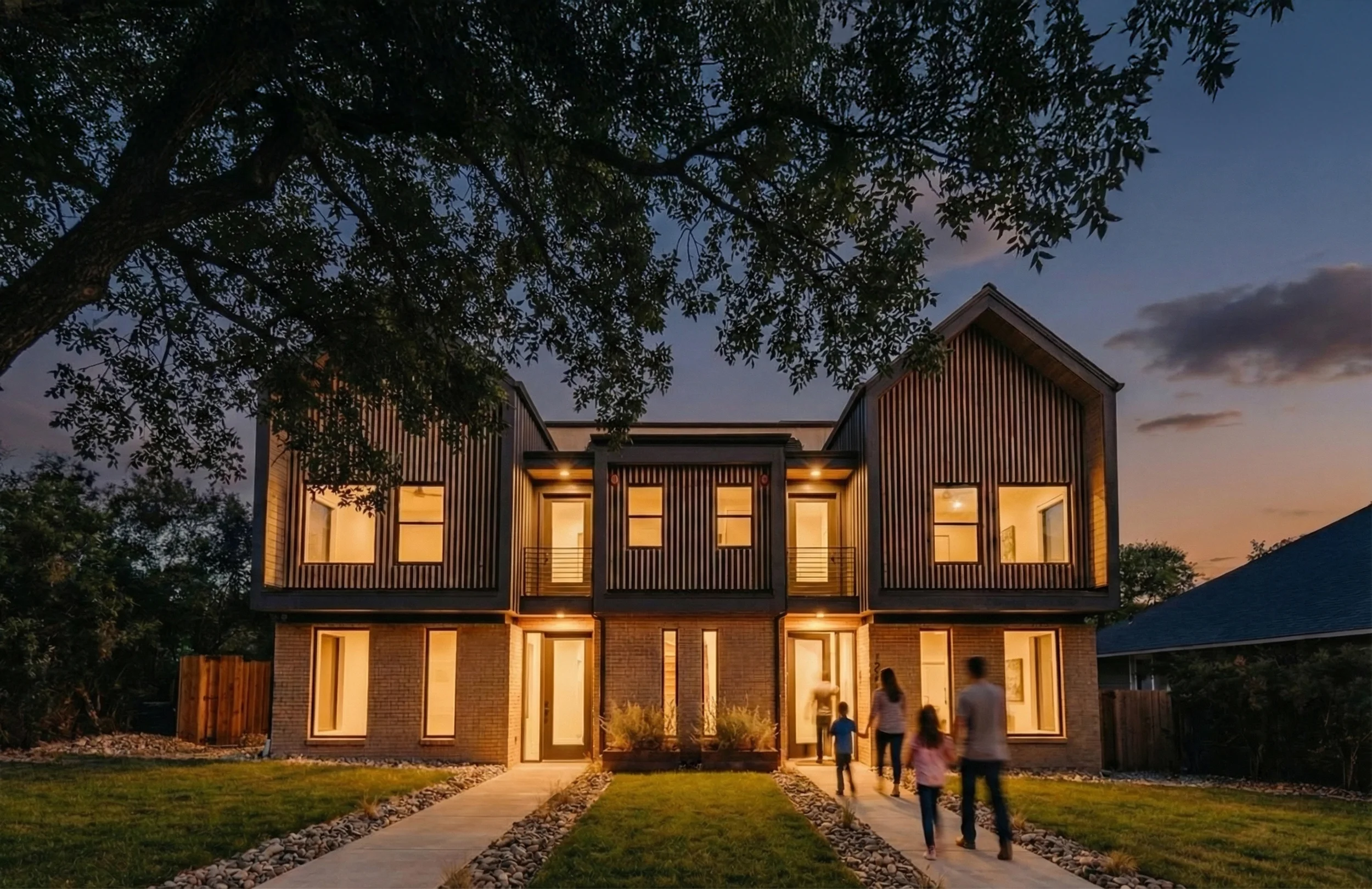 A modern two-story house with warm interior lights on an evening, featuring a brick lower level and black vertical siding on the upper level. A pathway leading to the front door is surrounded by grass and small rocks, with a family walking toward the house.