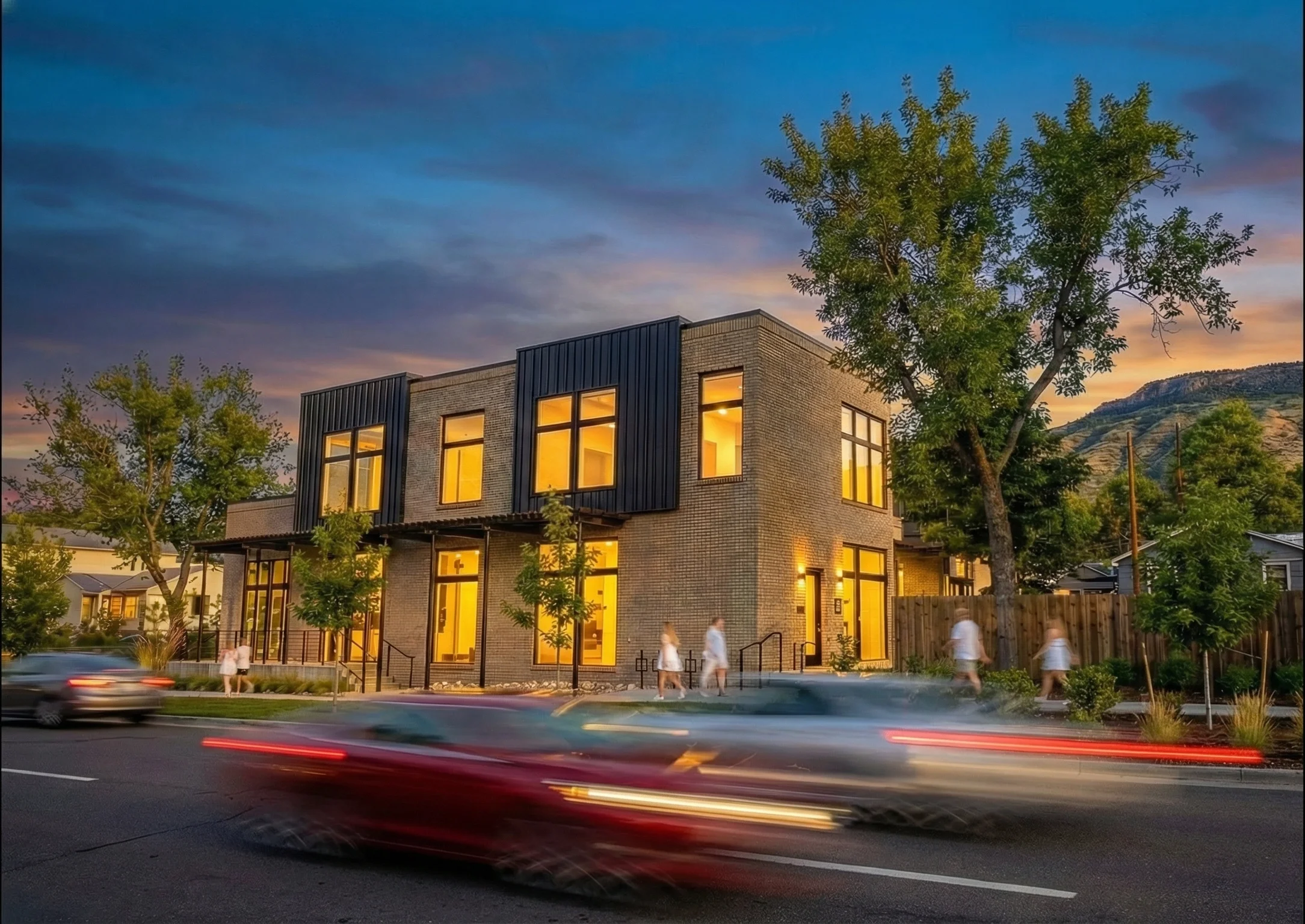 Modern brick building with large illuminated windows, trees in front, and blurred cars passing by on the street at sunset.