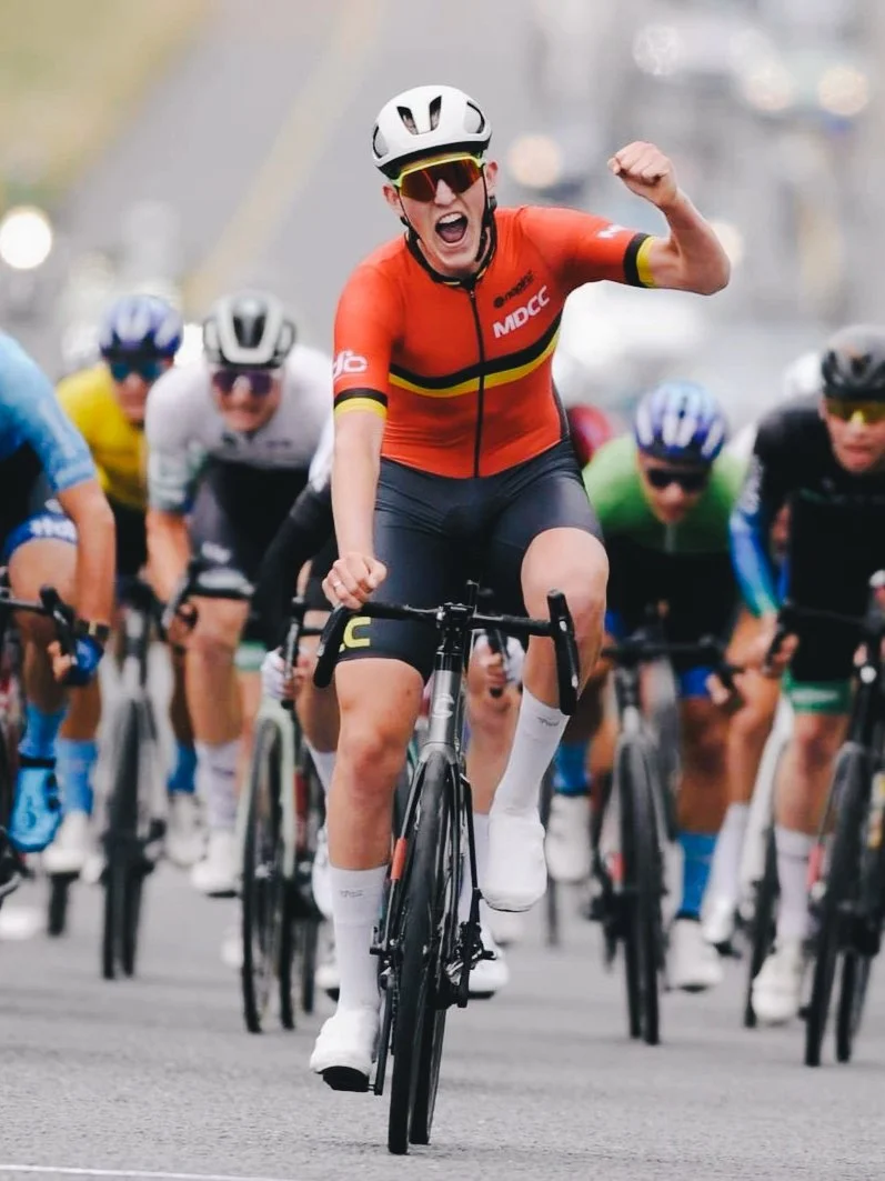 A male cyclist wearing a red jersey, black shorts, helmet, and sunglasses, raising his fist in celebration while riding among other cyclists during a race.