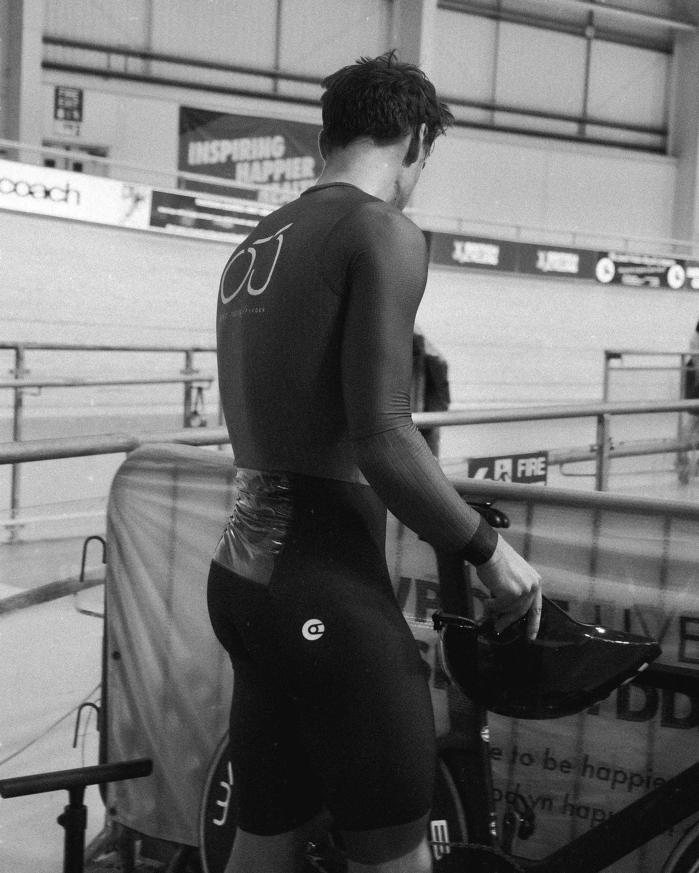 A male cyclist in a black long-sleeve cycling suit, holding a helmet, at an indoor velodrome.