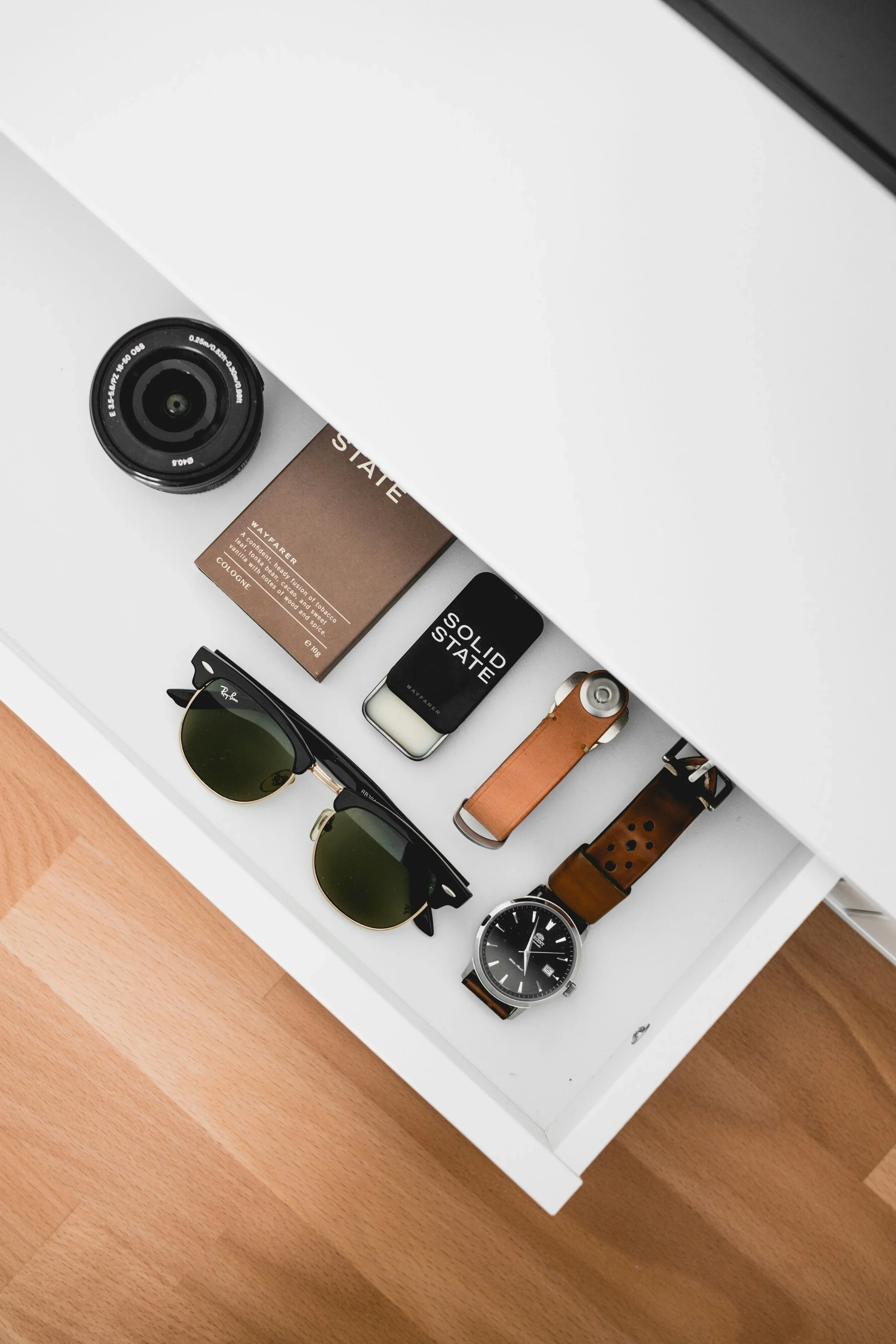 Organized drawer with neatly arranged sunglasses, watches, camera lens, and small accessories on a white surface.