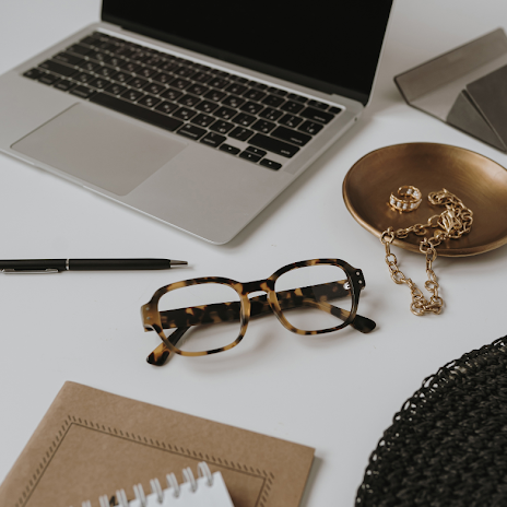 Minimal workspace with a laptop, tortoiseshell glasses, notebook, pen, and gold jewelry on a clean desk.