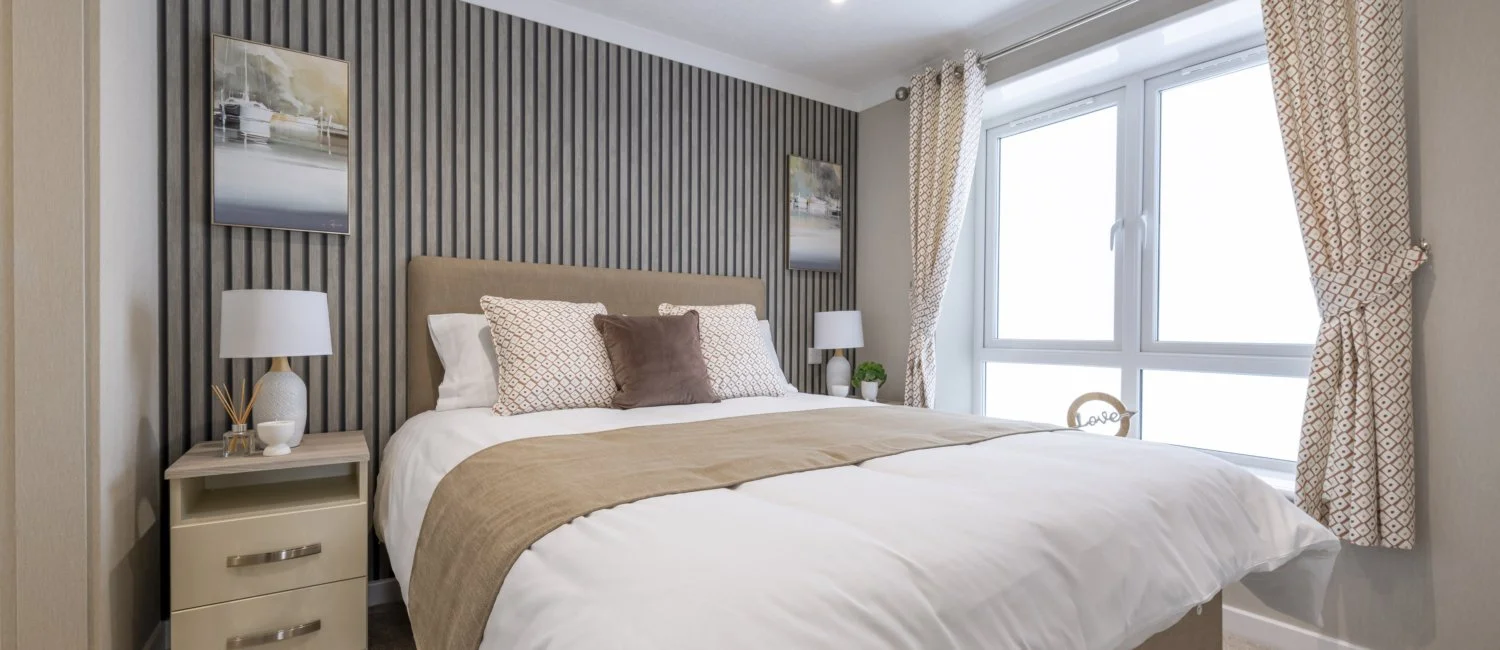 Bedroom with a large bed with beige headboard, white linens, and decorative pillows, flanked by bedside tables with lamps, patterned curtains, and large windows letting in natural light.