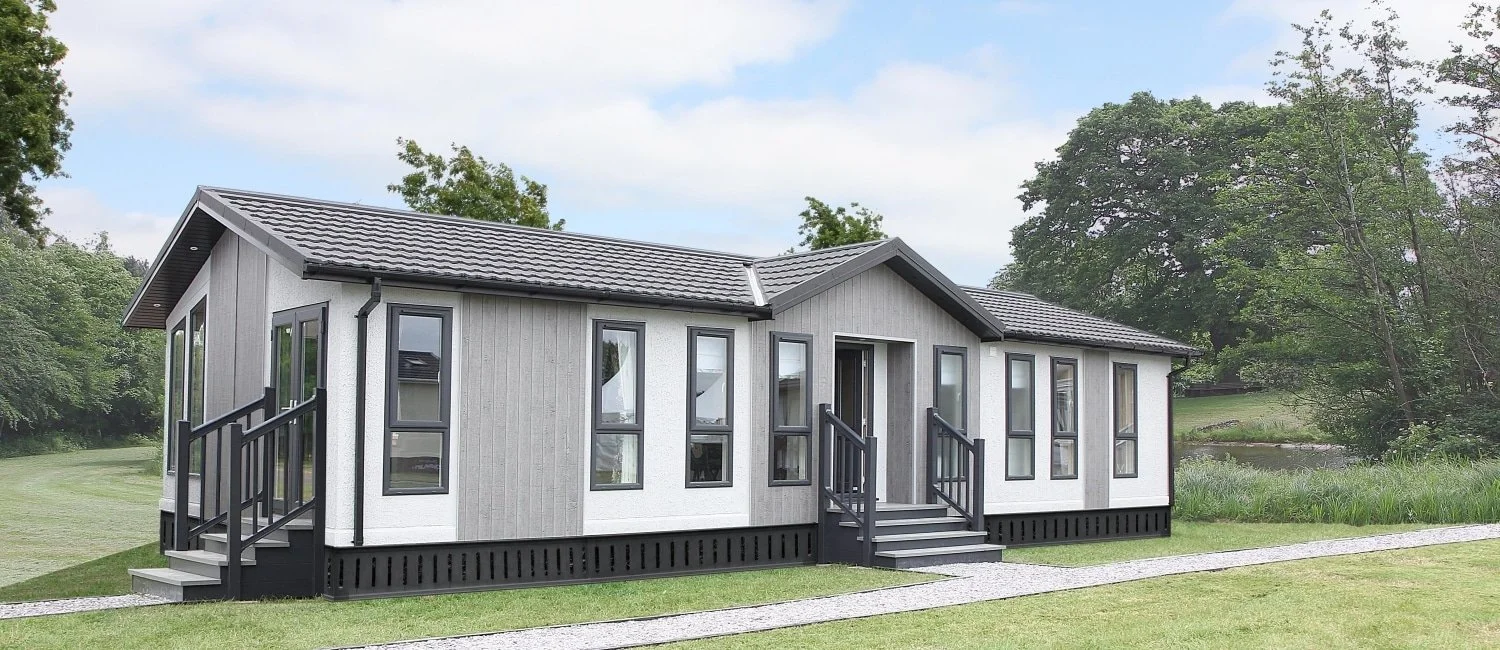 Modern tiny house with gray and white exterior, black stairs and railing, surrounded by green grass and trees.