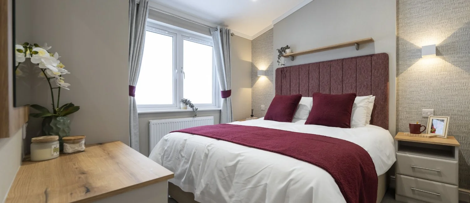 A cozy bedroom with a large bed featuring a burgundy headboard, pillows, and blanket, beside a window with curtains, with a nightstand, wall lamps, and decorative plants.