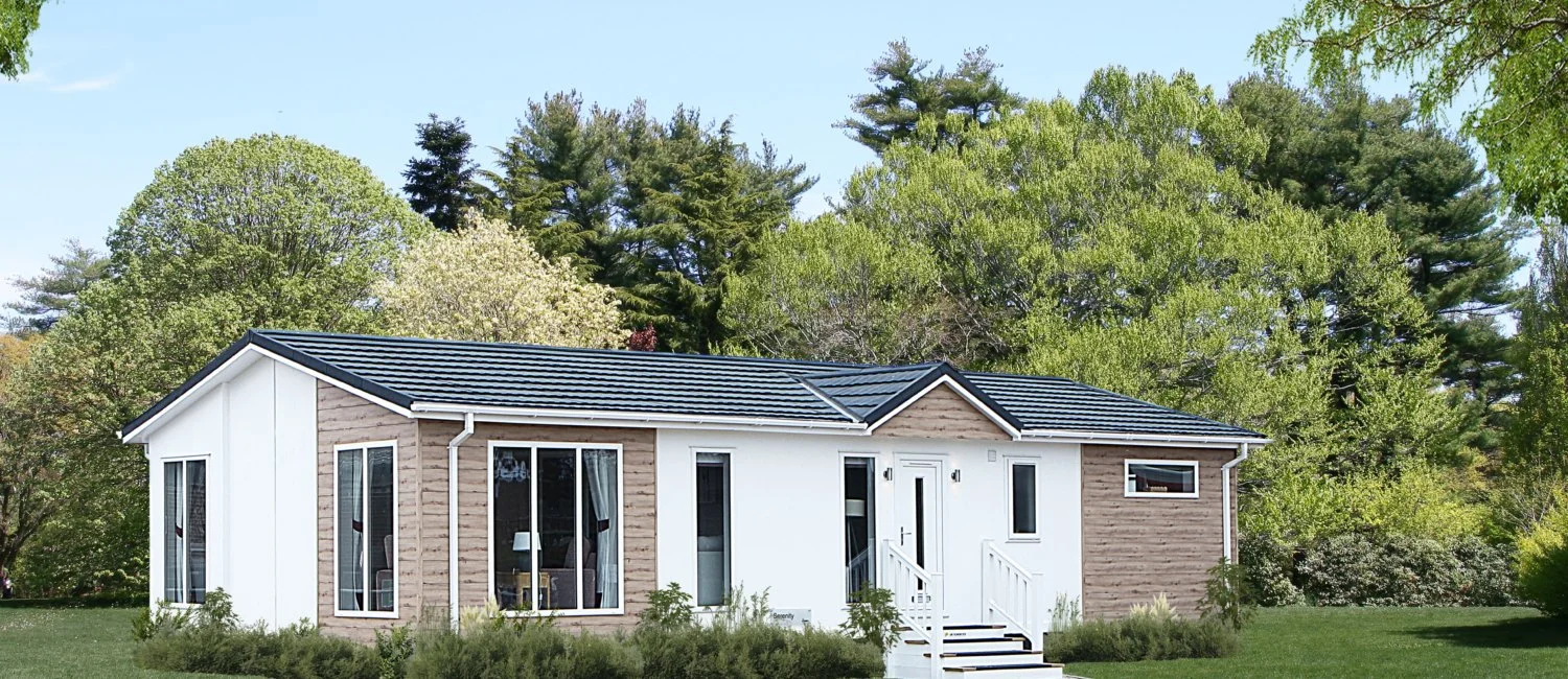 Modern single-story house with a black tile roof, white exterior walls, and wooden accents, situated in a lush green yard with trees in the background.