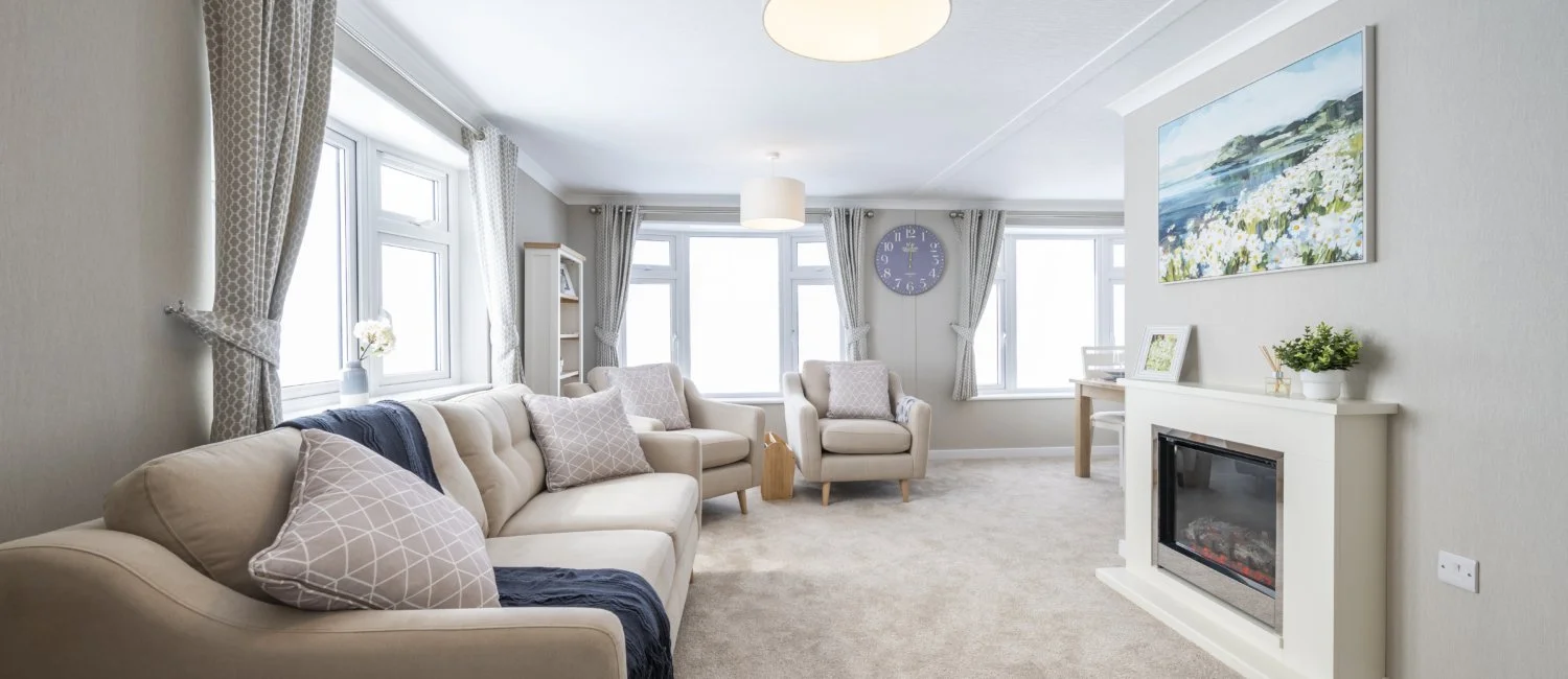 Bright living room with large bay windows, white furniture, a fireplace, a landscape painting, and decorative cushions.