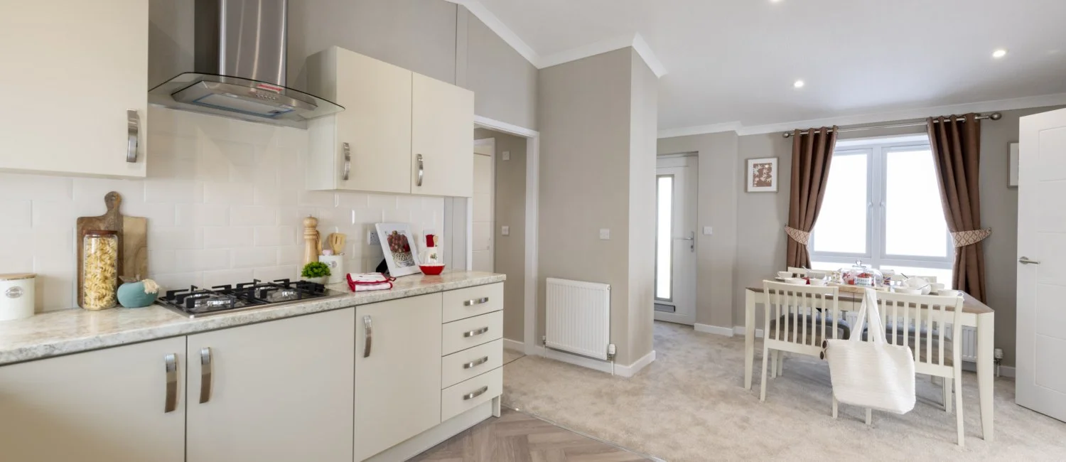 A modern kitchen and dining area with beige cabinetry, a gas stove, a marble countertop, and a white dining table with matching chairs, curtains with tiebacks, and a window with natural light.