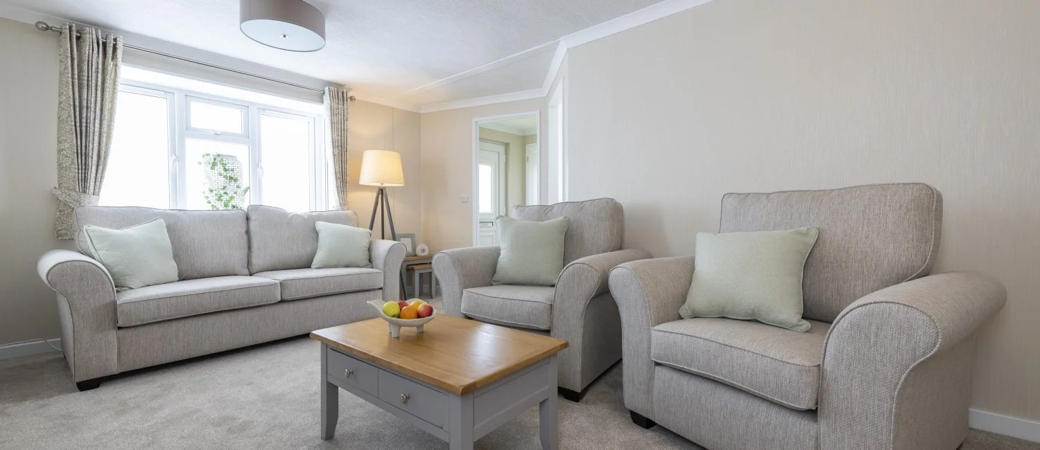 Living room with a beige sofa and two armchairs, a wooden coffee table with a bowl of fruit, a lamp, and a window with curtains.