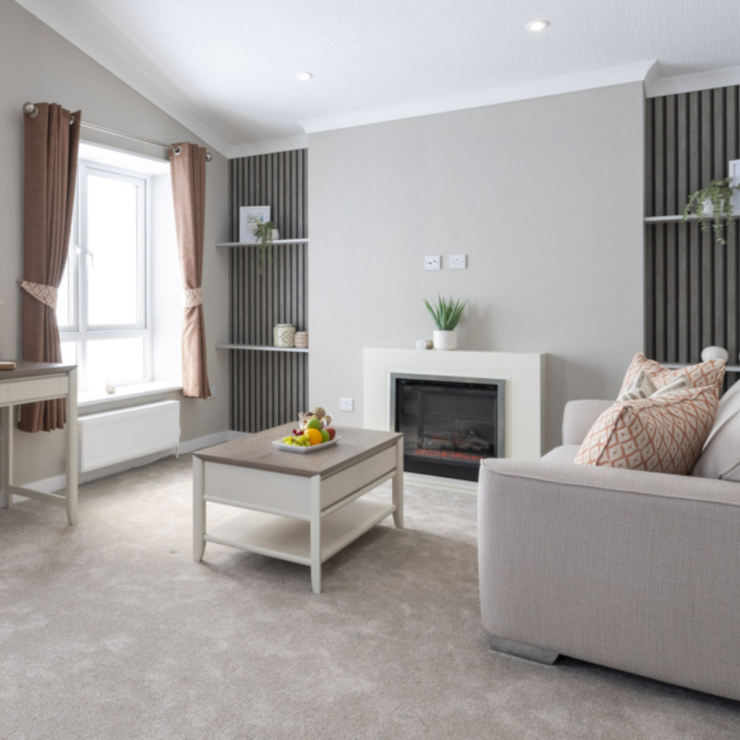Bright living room with beige carpet, white walls, and a large window with brown curtains. Contains a white sofa with patterned pillows, a gray and white coffee table with fruit, and a modern fireplace with potted plants on the mantle.