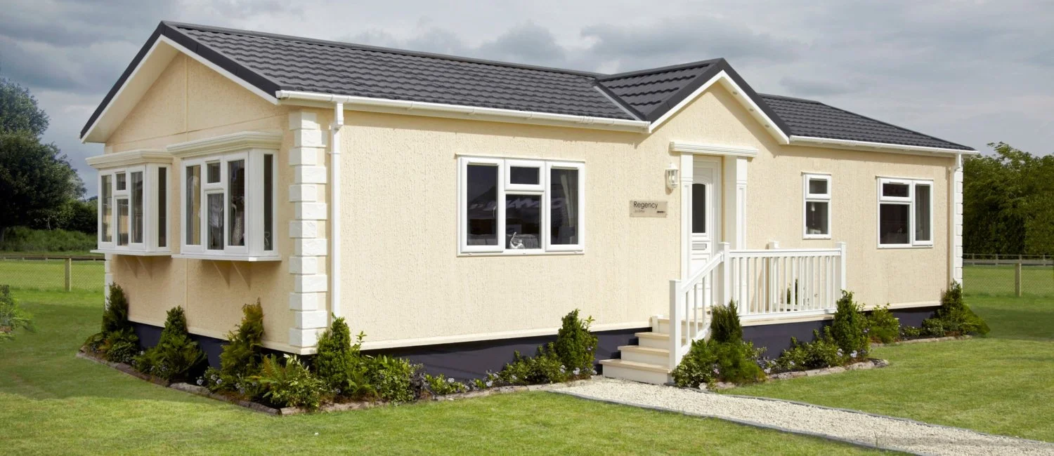 A beige single-story house with white trim, protected by a dark gray roof, surrounded by a green lawn and small shrubs near the foundation.