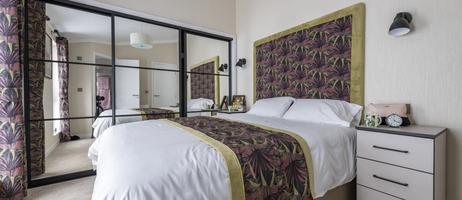 Bedroom with white bed, floral headboard and matching runner, mirrored wardrobe, nightstand with decor, and patterned curtains.