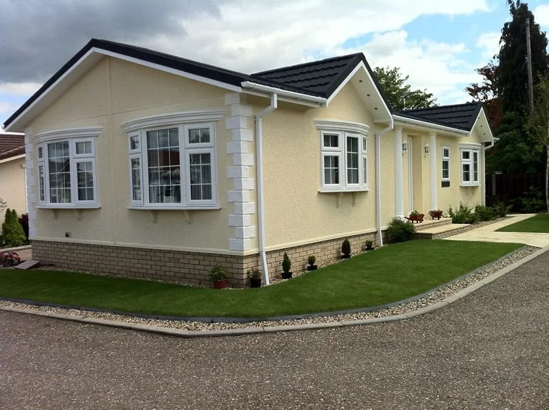 A yellow single-story house with white trim and black roof shingles, featuring bay windows, a front porch, and a well-maintained lawn with small bushes and potted plants.