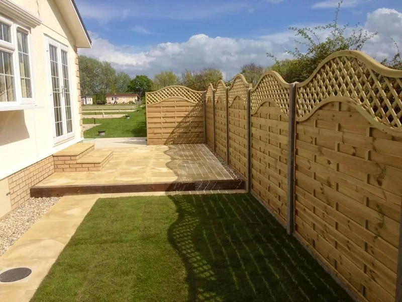 Newly installed wooden privacy fence along a yard with a grass lawn, patio, and steps leading to the porch of a house with large windows.