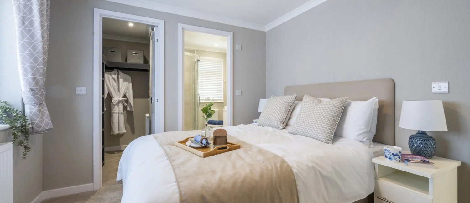A cozy bedroom with a bed, nightstand, and lamps. A tray with a journal and toiletries is on the bed. An open closet and an en-suite bathroom are visible.