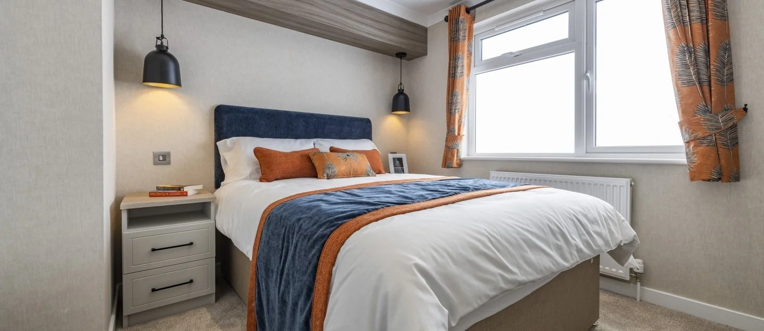 A neatly made bed with white sheets, an orange pillow, and a navy blue throw blanket in a bedroom. There is a gray nightstand with books and a framed photo next to the bed. Two black wall-mounted lamps hang above the bed. A large window with orange patterned curtains is on the right, allowing natural light into the room.