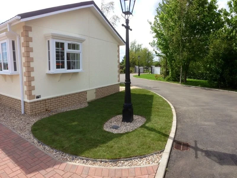 A curved driveway with a lamp post on a small patch of grass next to a cream-colored house with white-framed windows, bordered by gravel and brick pathway, and surrounded by green trees.