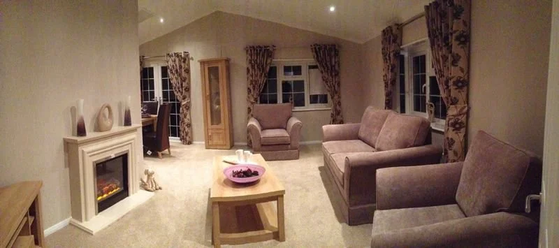 Living room with beige walls and carpet, purple sofas, a wooden coffee table with a pink bowl, patterned curtains, a fireplace, and a corner cabinet.