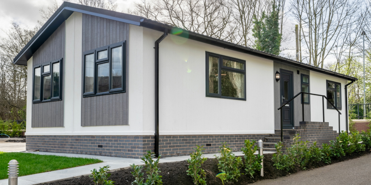 A modern, single-story house with a combination of white and gray exterior, black window frames, a black door, brick stairs, and landscaping with green bushes.