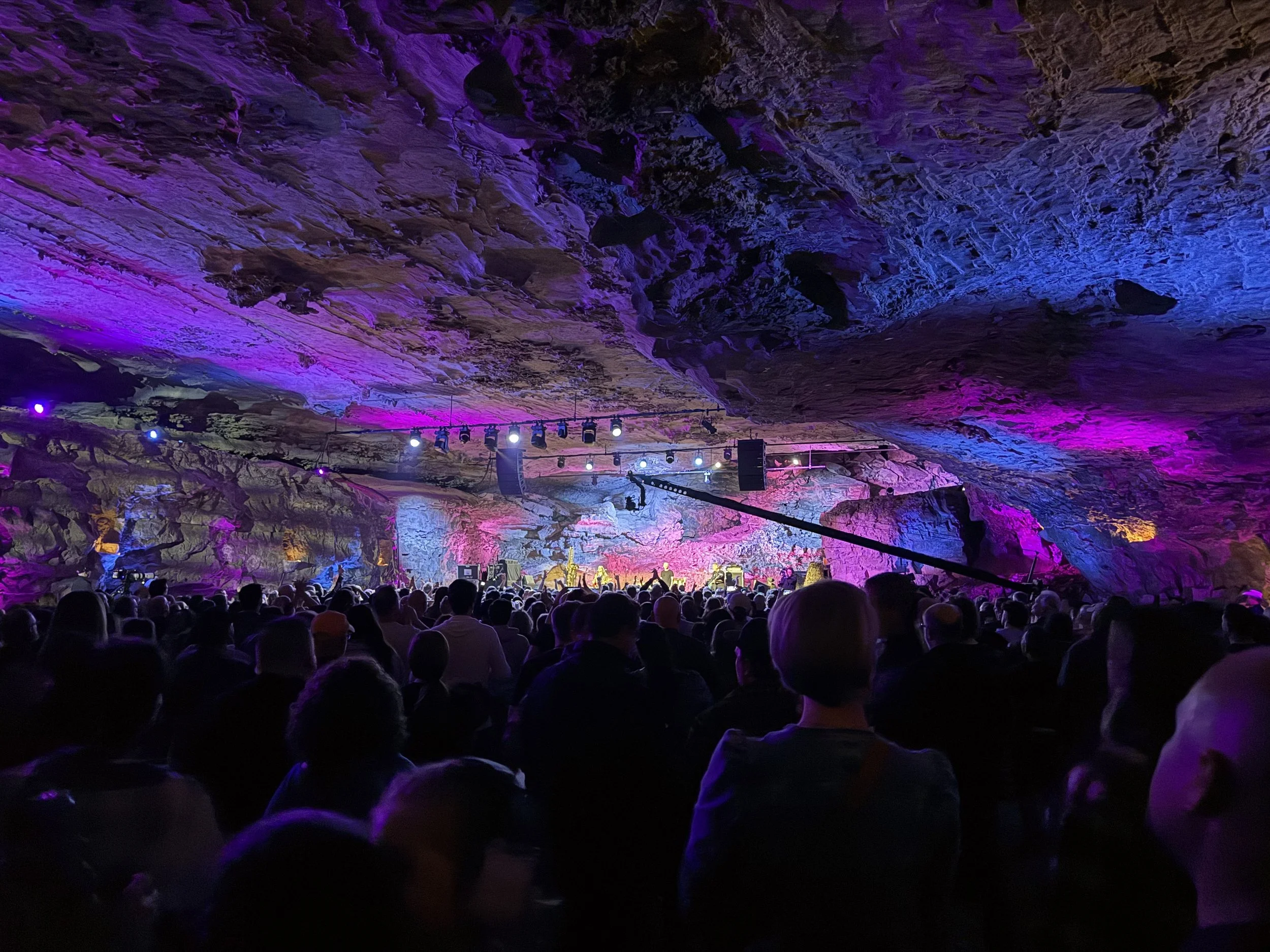 An audience watching a concert inside a large cavern with rocky walls illuminated by colorful lights.