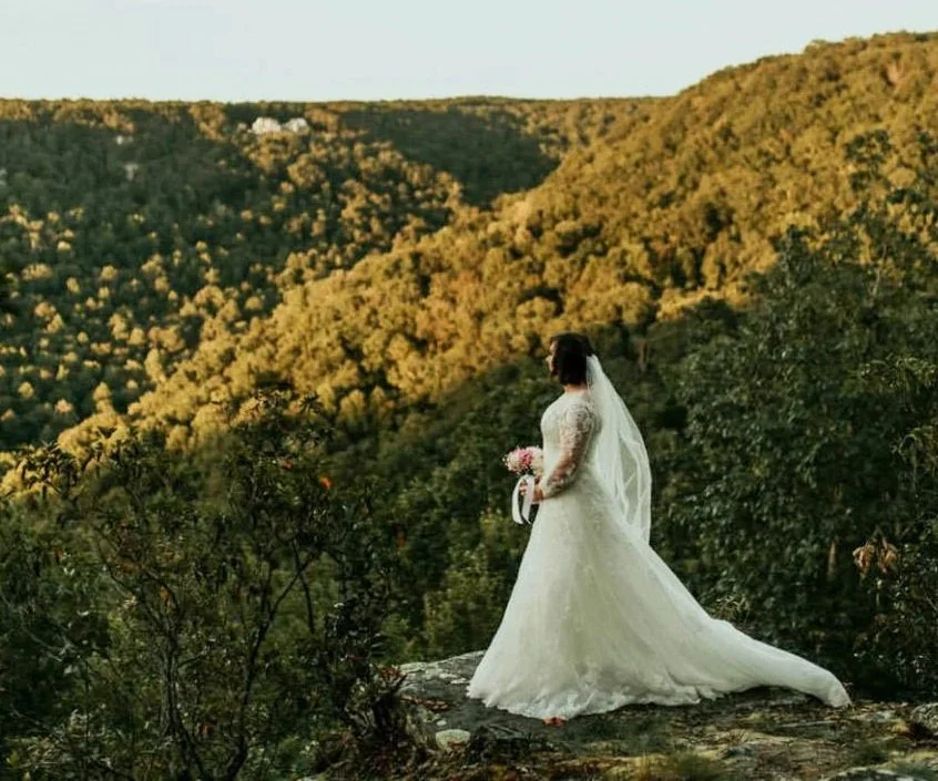 A bride in a white wedding gown holding a bouquet, standing on a rocky ledge amid green trees and overlooking a hilly landscape during sunset.