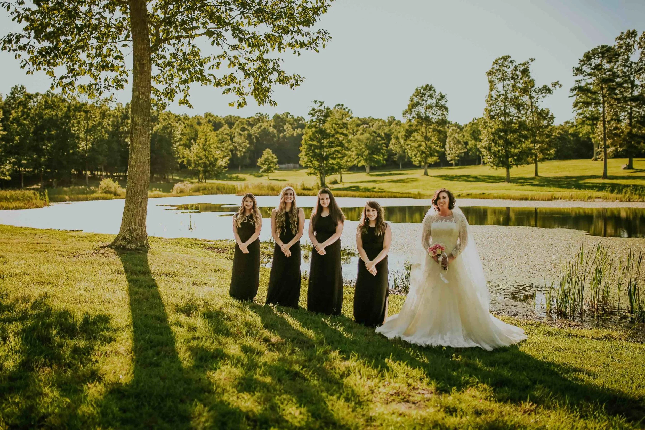A bride in a white wedding dress holding a bouquet and four bridesmaids in black dresses standing by a pond in a green park with trees, on a sunny day.