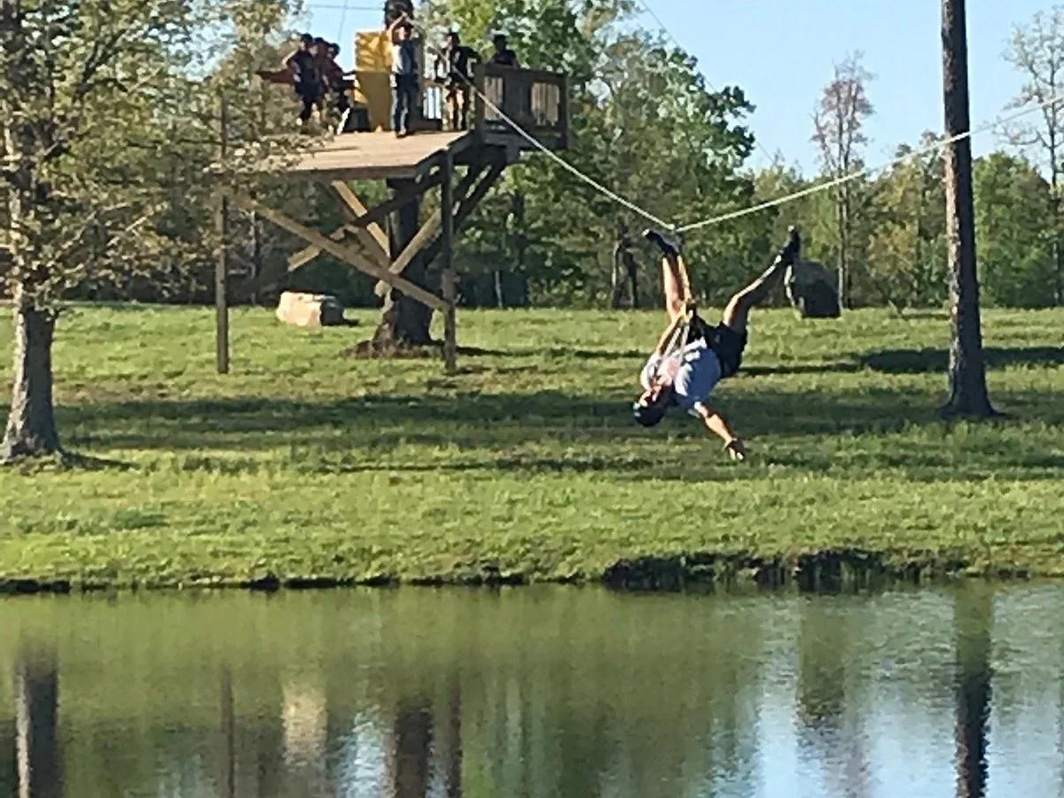 Person swinging upside down on a rope over a pond at an outdoor park with trees and a wooden deck in the background.
