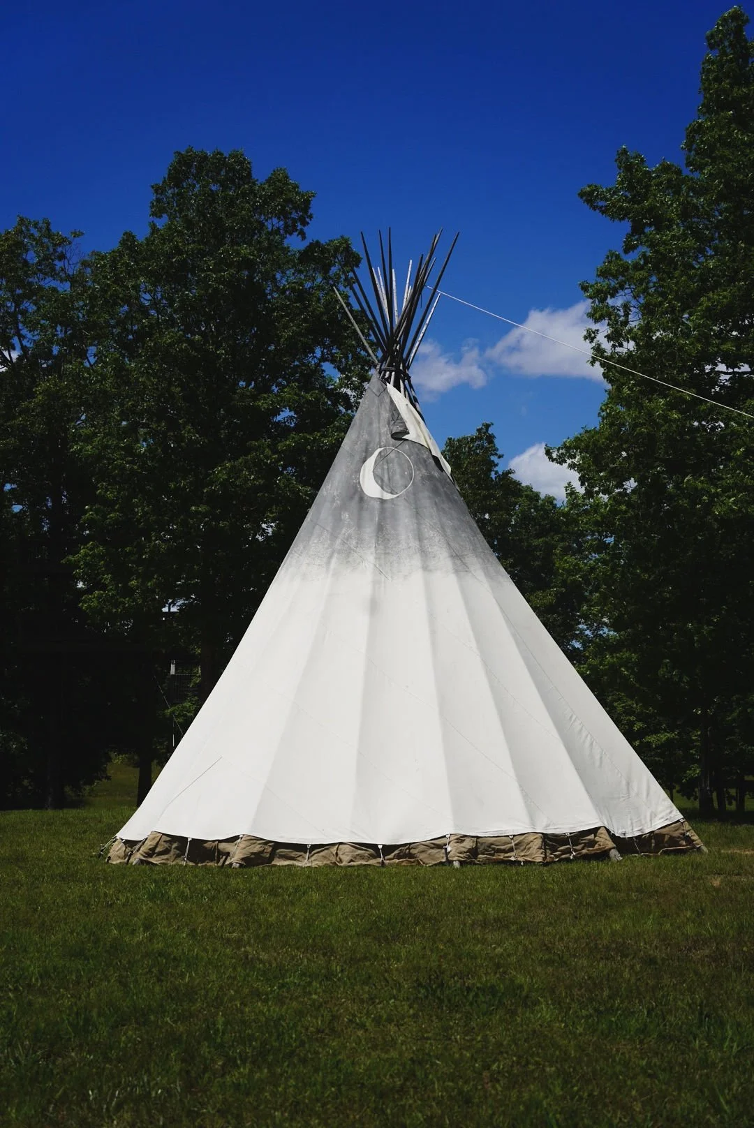 A white teepee tent set up on a grassy area with trees and a blue sky with some clouds in the background.